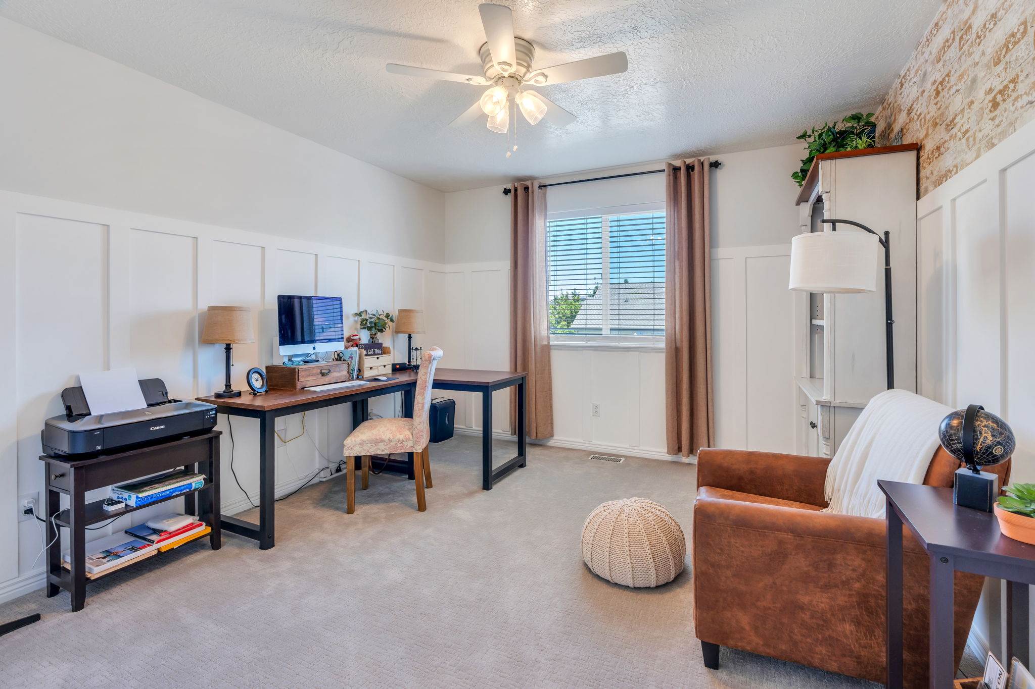 Board and batten wainscoting frames this versatile room, featuring a window with blinds, ceiling fan, and neutral carpet flooring