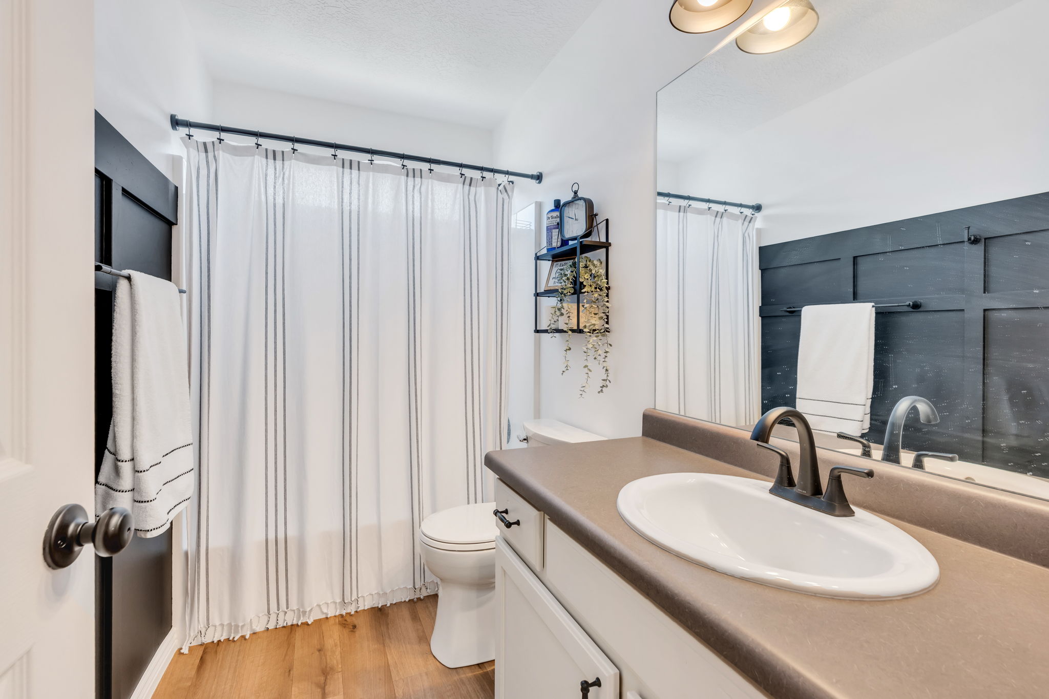 Bathroom featuring wood-finish flooring, a white vanity with dark hardware, a single basin sink with a dark bronze faucet, and a dark accent wall with board and batten detailing