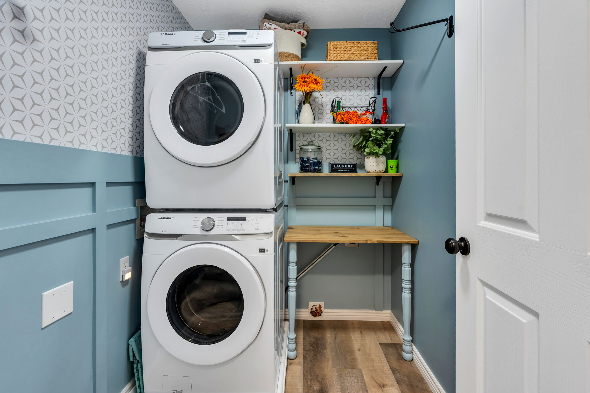 Dedicated laundry area featuring a stacked washer and dryer, wood-finish flooring, and a folding table with shelving