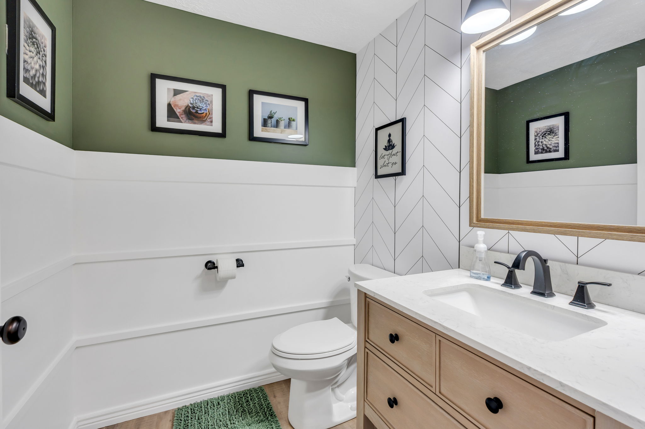 Powder room featuring a light wood-finish vanity with a built-in sink, dark bronze fixtures, a framed mirror, and a chevron tile accent wall