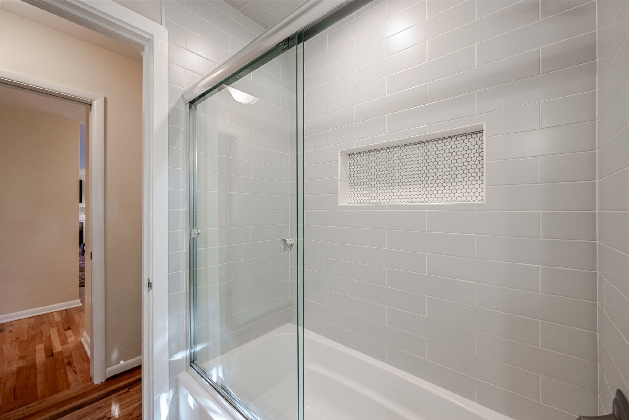 Bathtub with sliding glass enclosure, light gray subway tile surround, and a recessed niche