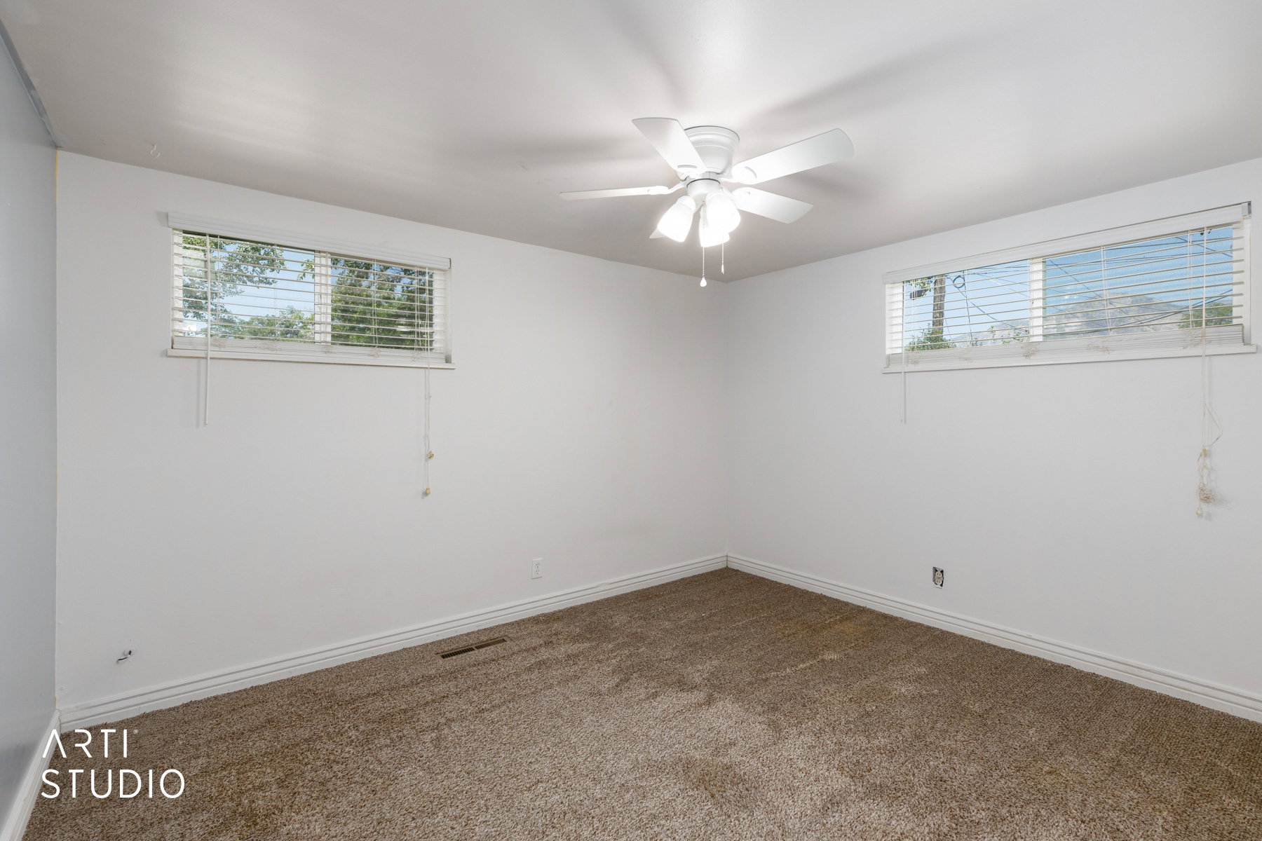 Neutral wall tones complementing plush carpeting, featuring twin windows with horizontal blinds, a ceiling fan, and baseboard trim