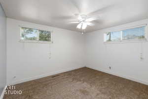 Neutral wall tones complementing plush carpeting, featuring twin windows with horizontal blinds, a ceiling fan, and baseboard trim
