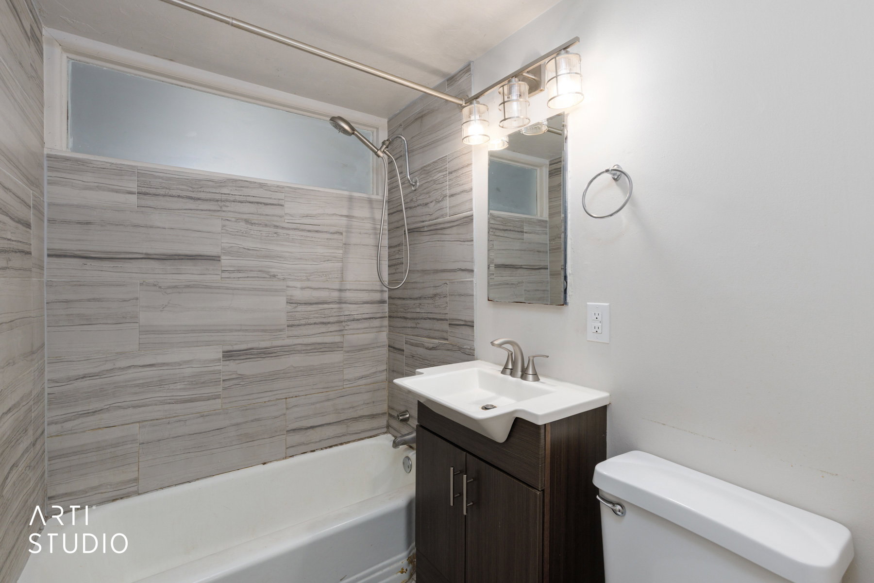 Bathroom featuring a white bathtub with a tiled shower surround, an opaque transom window, a dark wood-finish vanity with an integrated sink, a brushed nickel faucet, and a wall-mounted light fixture