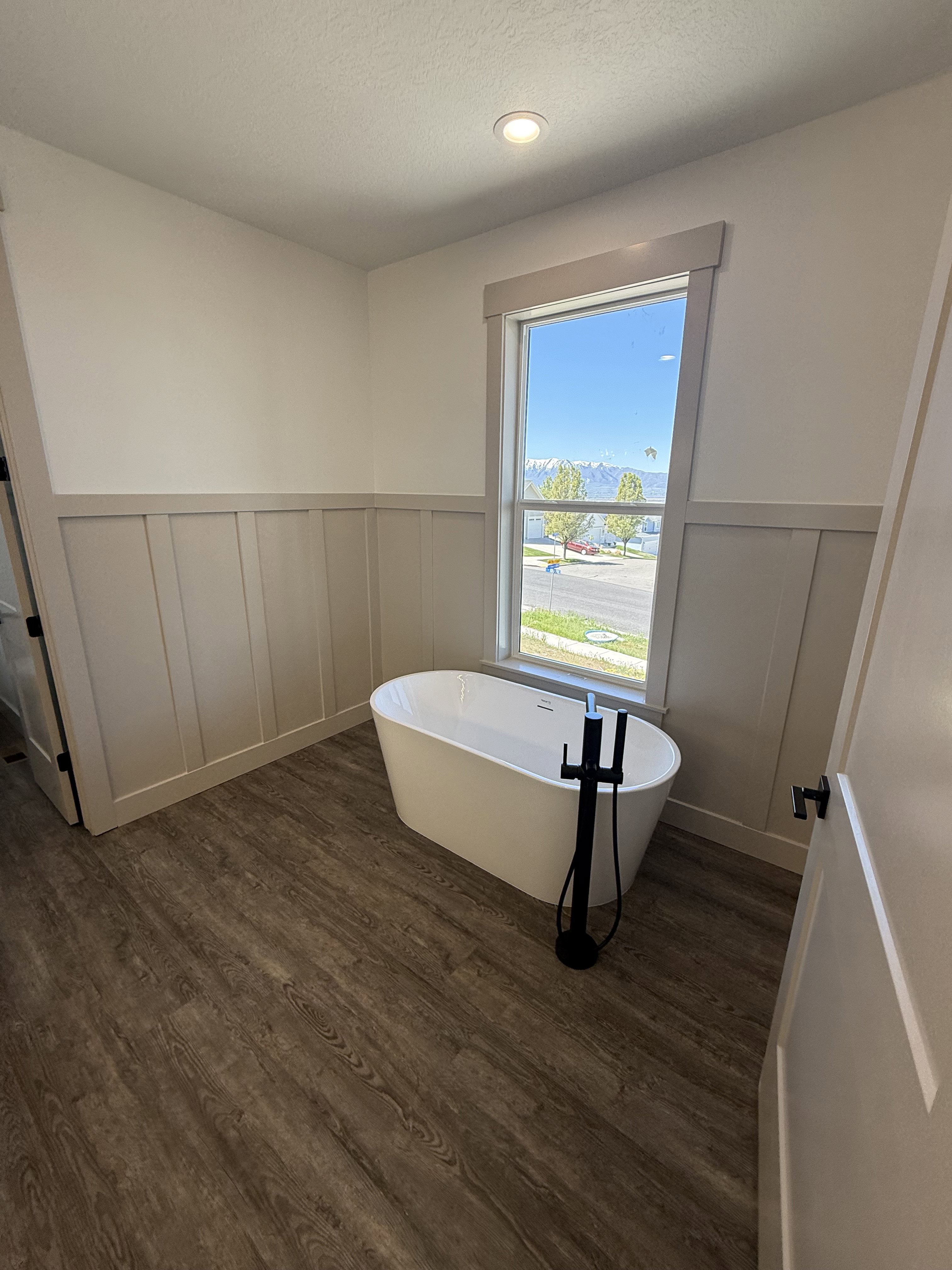 Freestanding soaking tub with a matte black floor-mounted faucet, wood-finish flooring, and wainscoting
