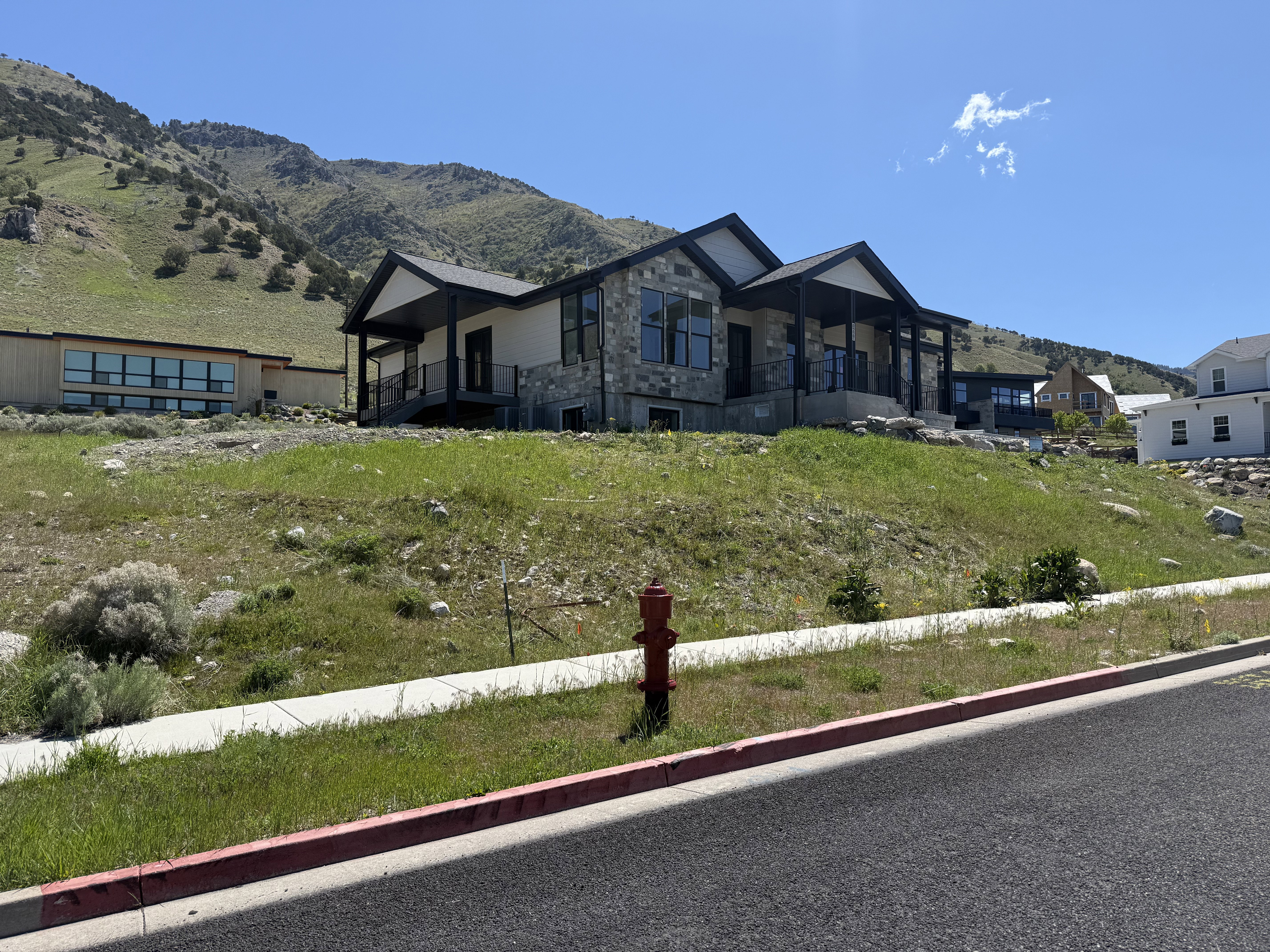 Modern mountain home featuring stone and siding exterior, large windows, an expansive covered porch with dark railings, and a hillside setting