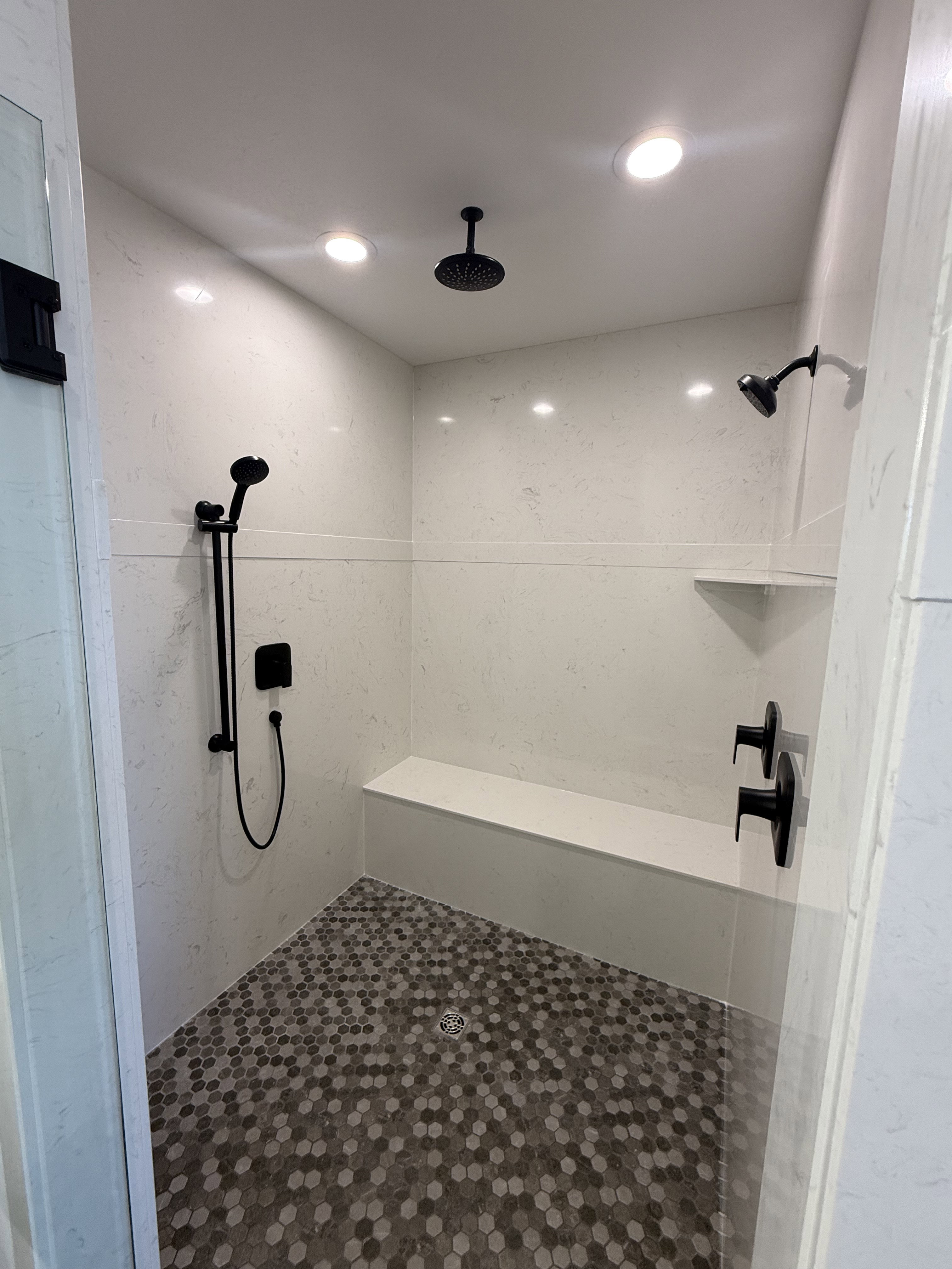 Spacious walk-in shower featuring a rain shower head, additional shower head with handheld sprayer, matte black fixtures, built-in bench, and hexagonal mosaic tile flooring