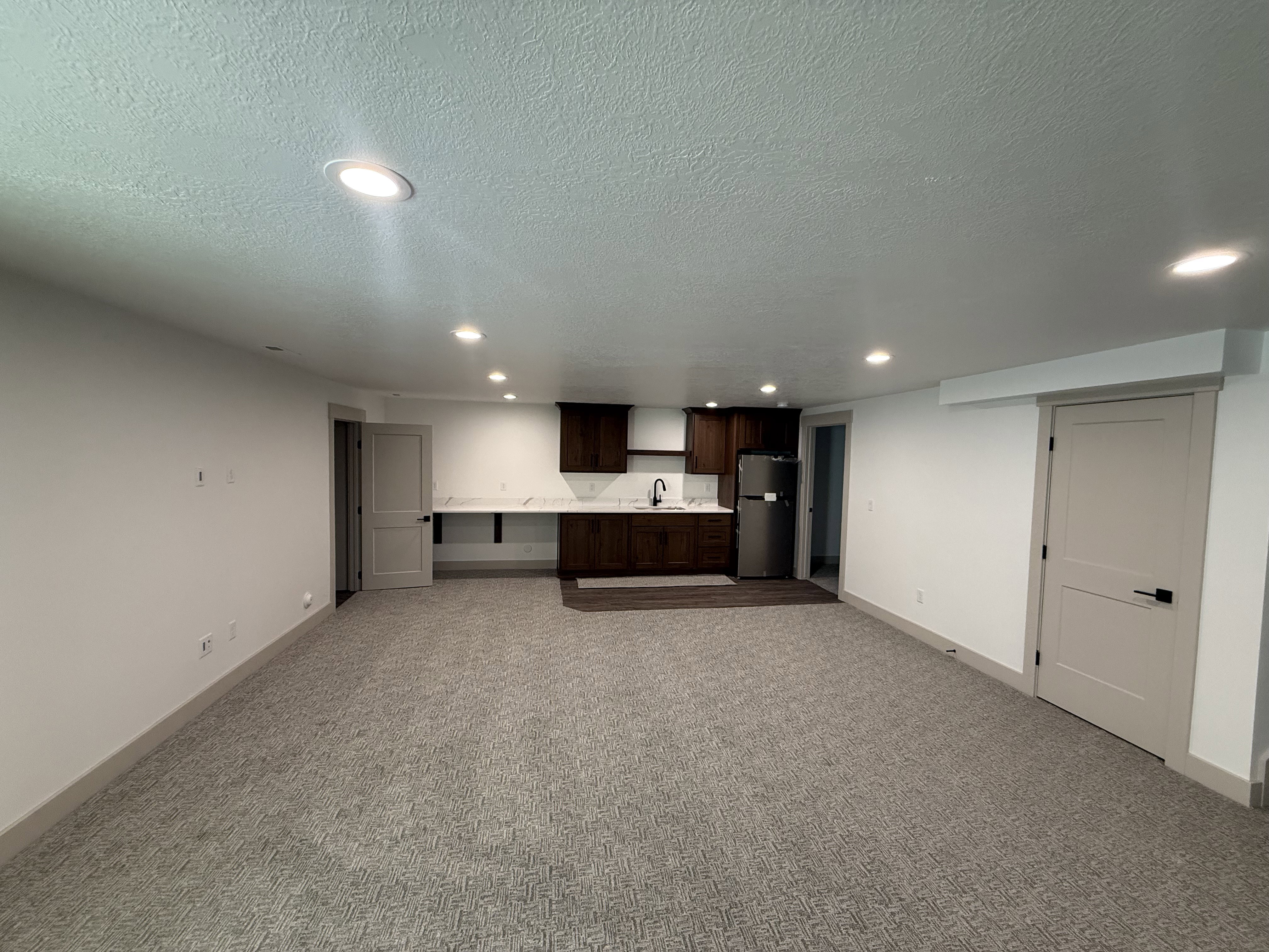 Spacious room featuring recessed lighting, a built-in kitchenette with dark wood-finish cabinetry, a stainless steel refrigerator, and a white countertop with an undermount sink