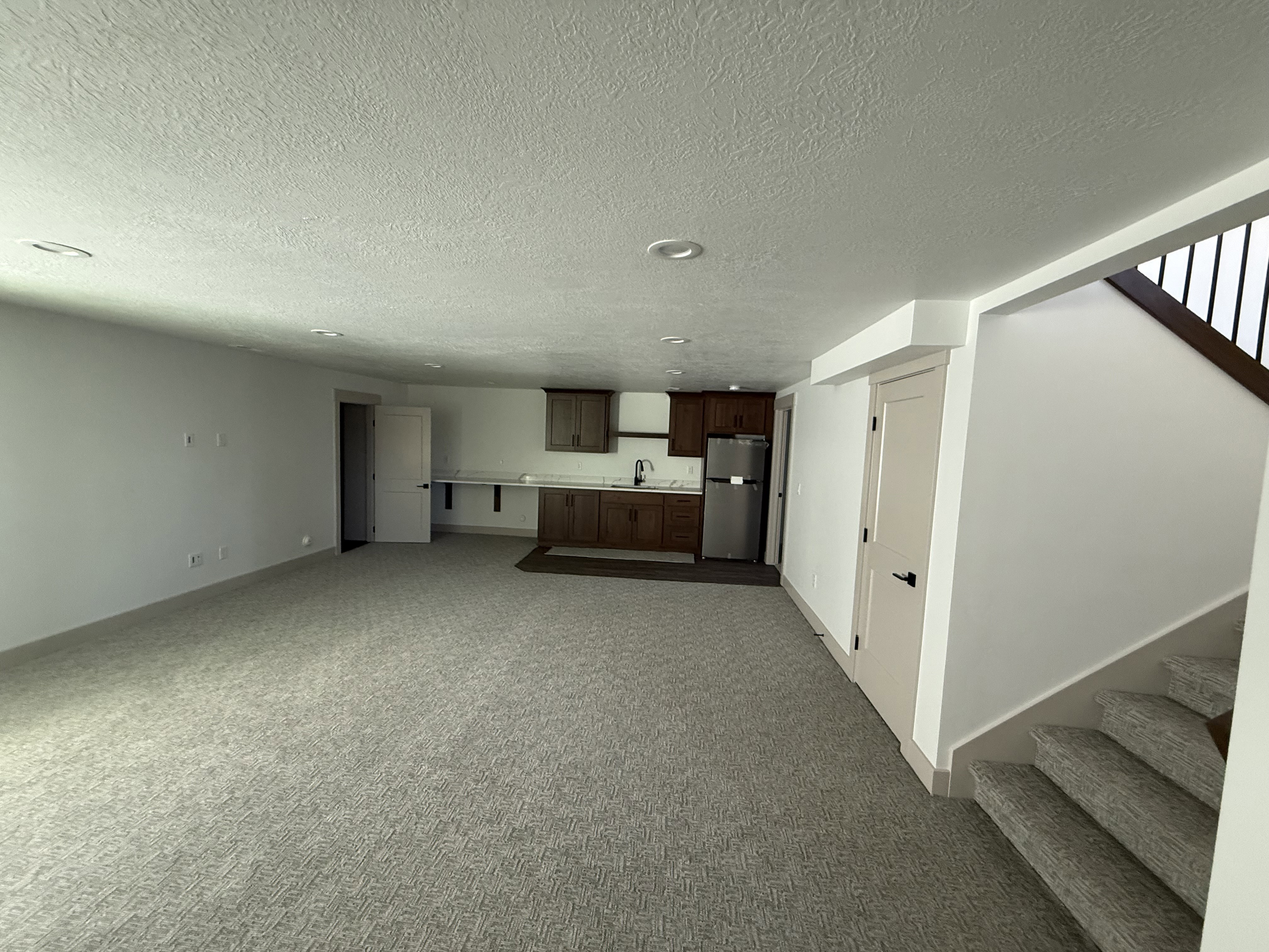 Spacious lower-level area featuring plush carpet flooring, recessed lighting, and a kitchenette with wood cabinetry, a stainless steel refrigerator, and a sink