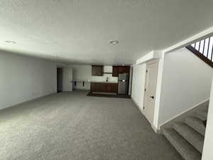 Spacious lower-level area featuring plush carpet flooring, recessed lighting, and a kitchenette with wood cabinetry, a stainless steel refrigerator, and a sink