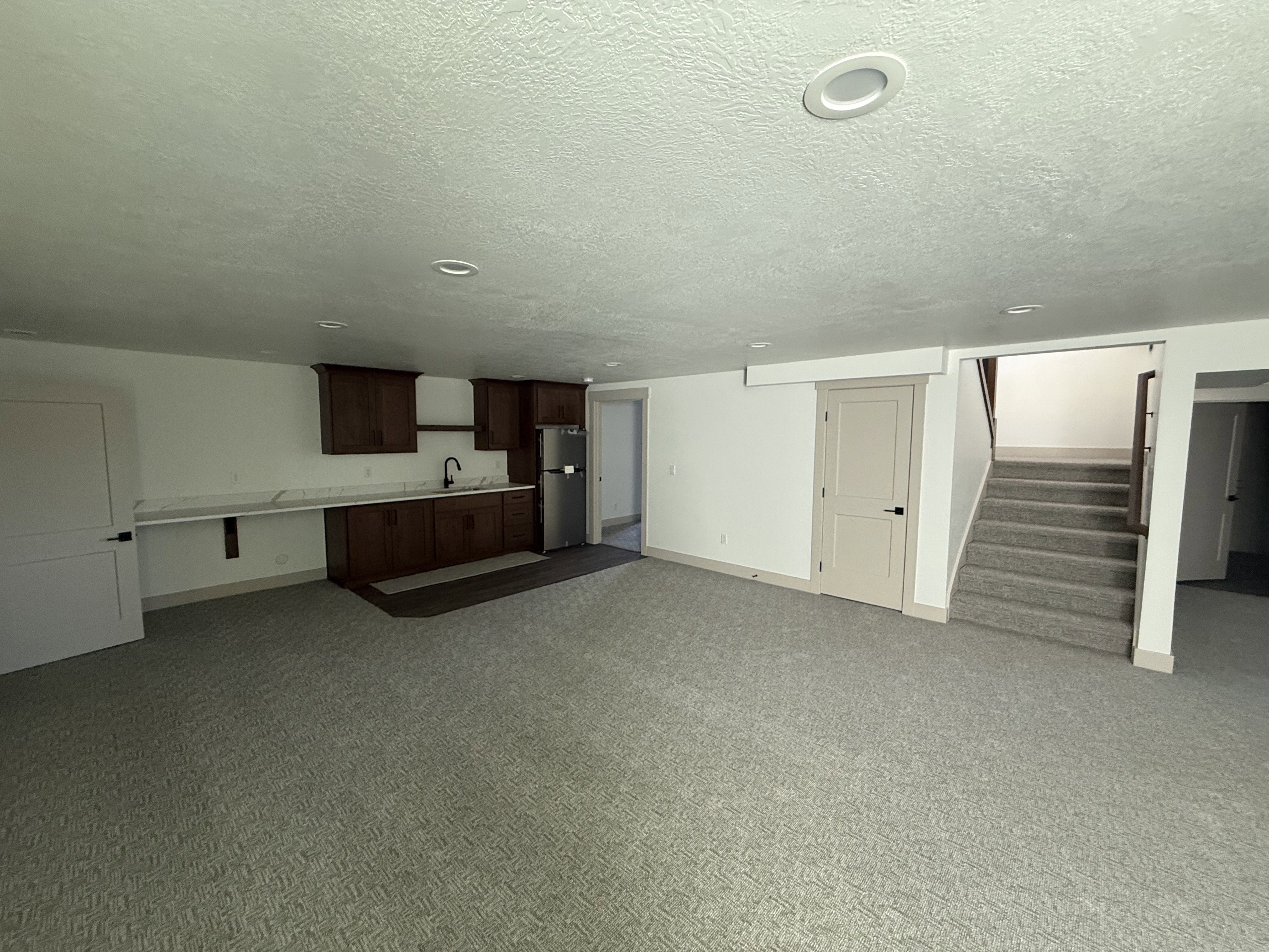 Spacious lower-level area featuring a kitchenette with dark wood cabinetry, a stainless steel refrigerator, and a sink with a matte black faucet