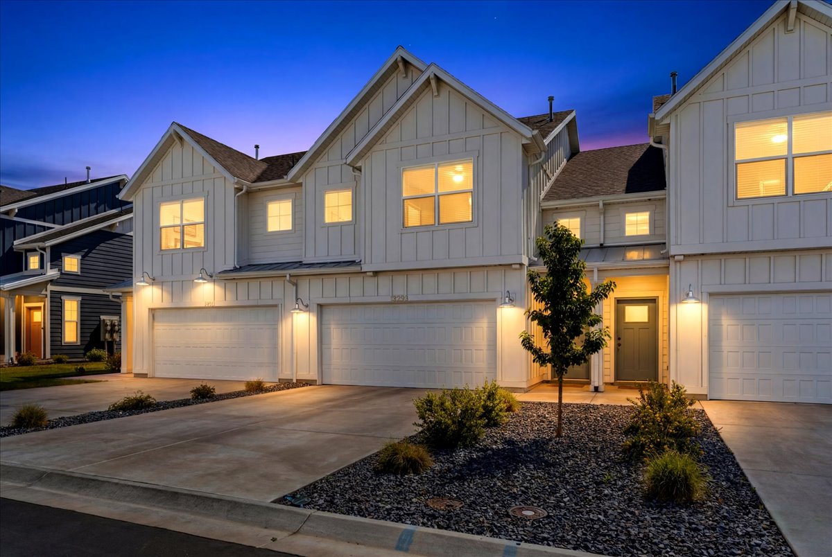Modern farmhouse-style townhome featuring white board and batten siding, multiple windows, a dark gray front door with glass insert, and an attached two-car garage with overhead lighting