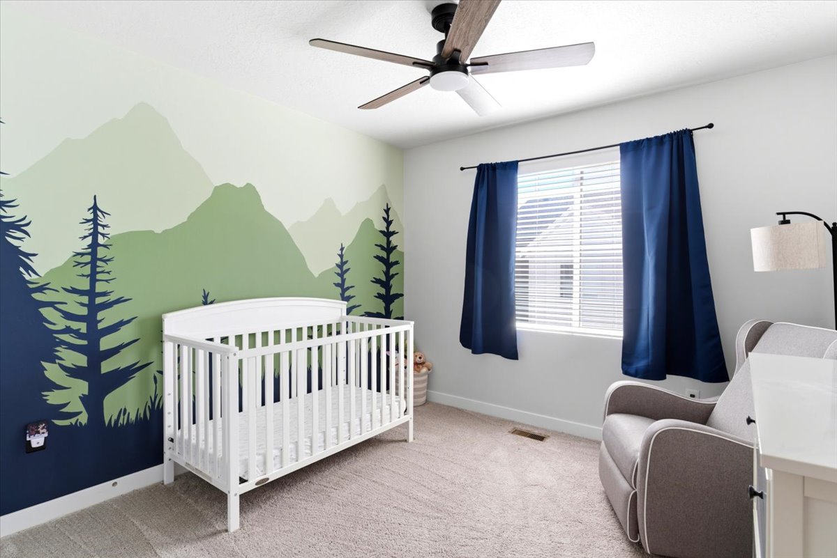 Light-colored carpeted flooring extending throughout, a ceiling fan with dark wood-finish blades, a window with horizontal blinds and dark blue drapes, and a wall featuring a painted mountain and tree mural