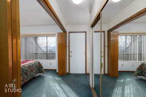 Bedroom featuring a large window with vertical blinds, teal carpet flooring, and a white entry door with a brass-finish knob