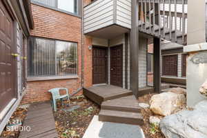 Exterior entrance featuring brick and horizontal siding, multiple brown paneled entry doors, an upper-level balcony with wood railing, and a ground-level wood walkway with steps