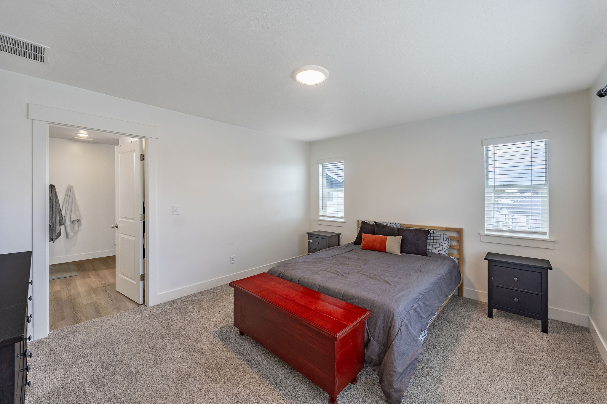 Spacious master bedroom featuring neutral carpeting, white walls, two windows with blinds, an en suite bathroom with wood-finish flooring, and a ceiling-mounted light fixture