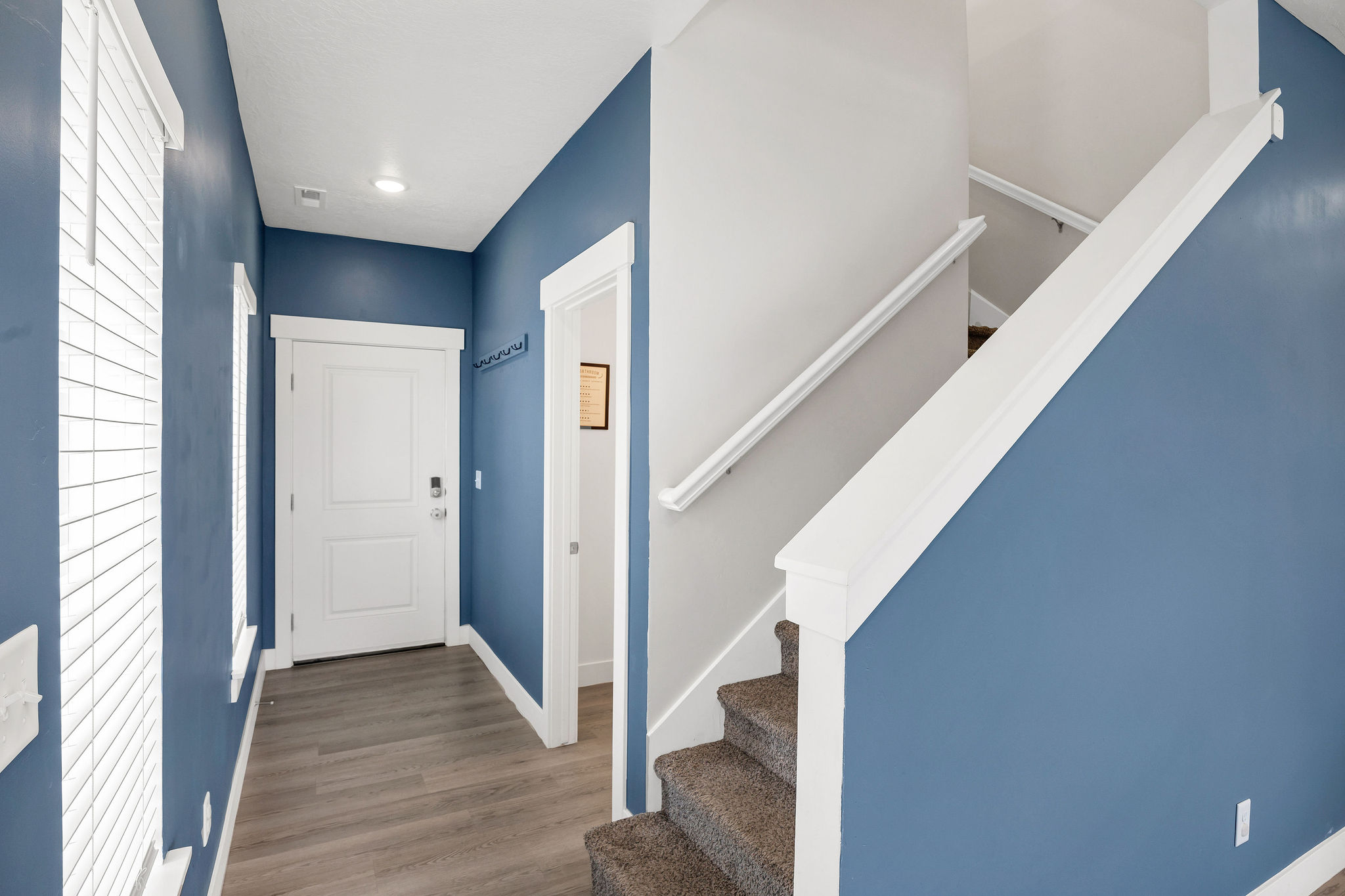 Entryway featuring wood-finish flooring, two windows with blinds, a white paneled door with smart lock hardware, and a staircase with carpeted treads and a white handrail