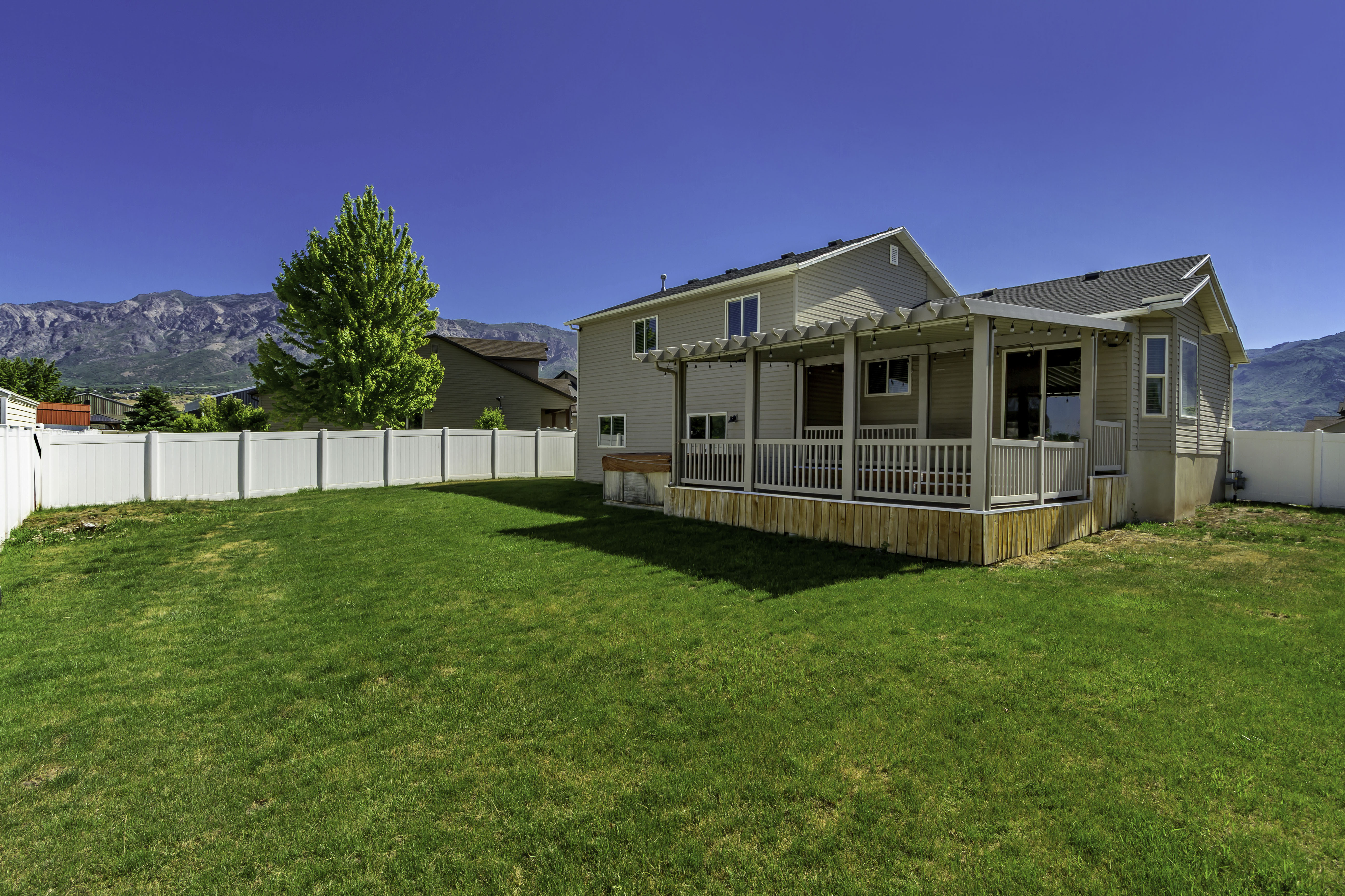 Spacious backyard featuring a manicured lawn, white vinyl fencing, a covered patio with a pergola, and mountain scenery in the distance