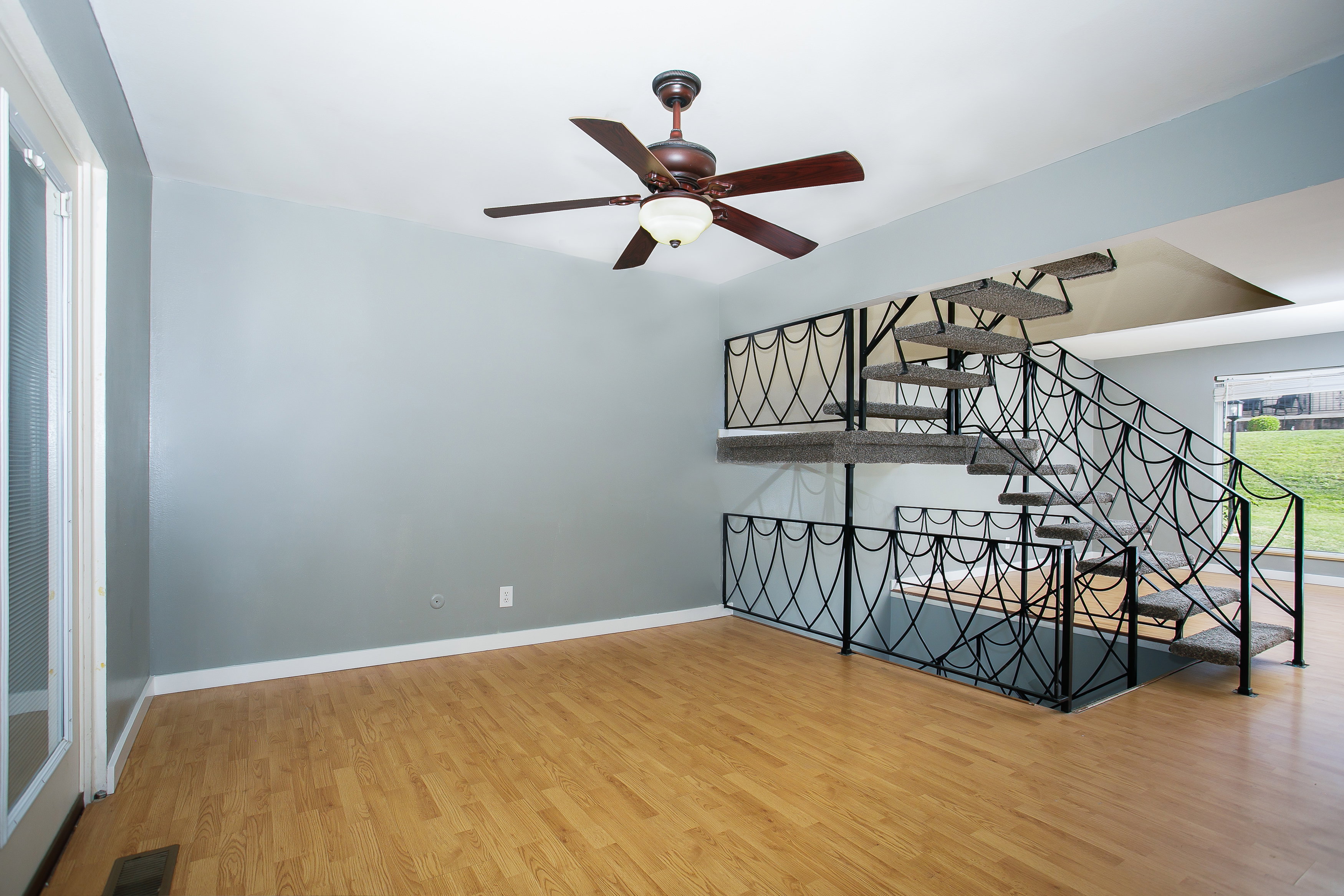 Spacious room featuring wood-finish flooring, a contemporary ceiling fan, and a striking black wrought iron staircase with carpeted treads