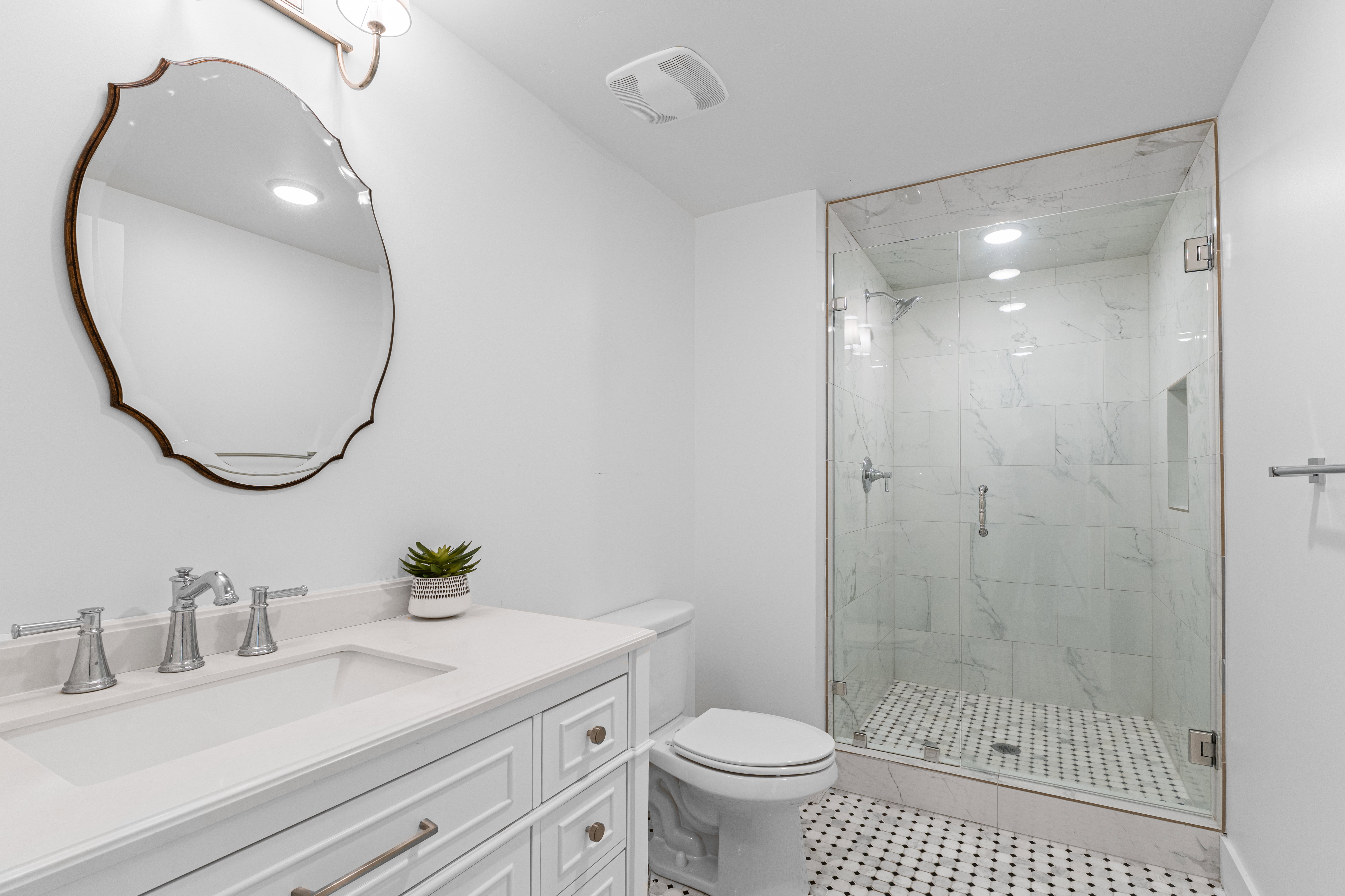 Bathroom featuring a frameless glass shower enclosure with marble-patterned tile, a white vanity with drawers, a single basin sink, and a decorative framed mirror