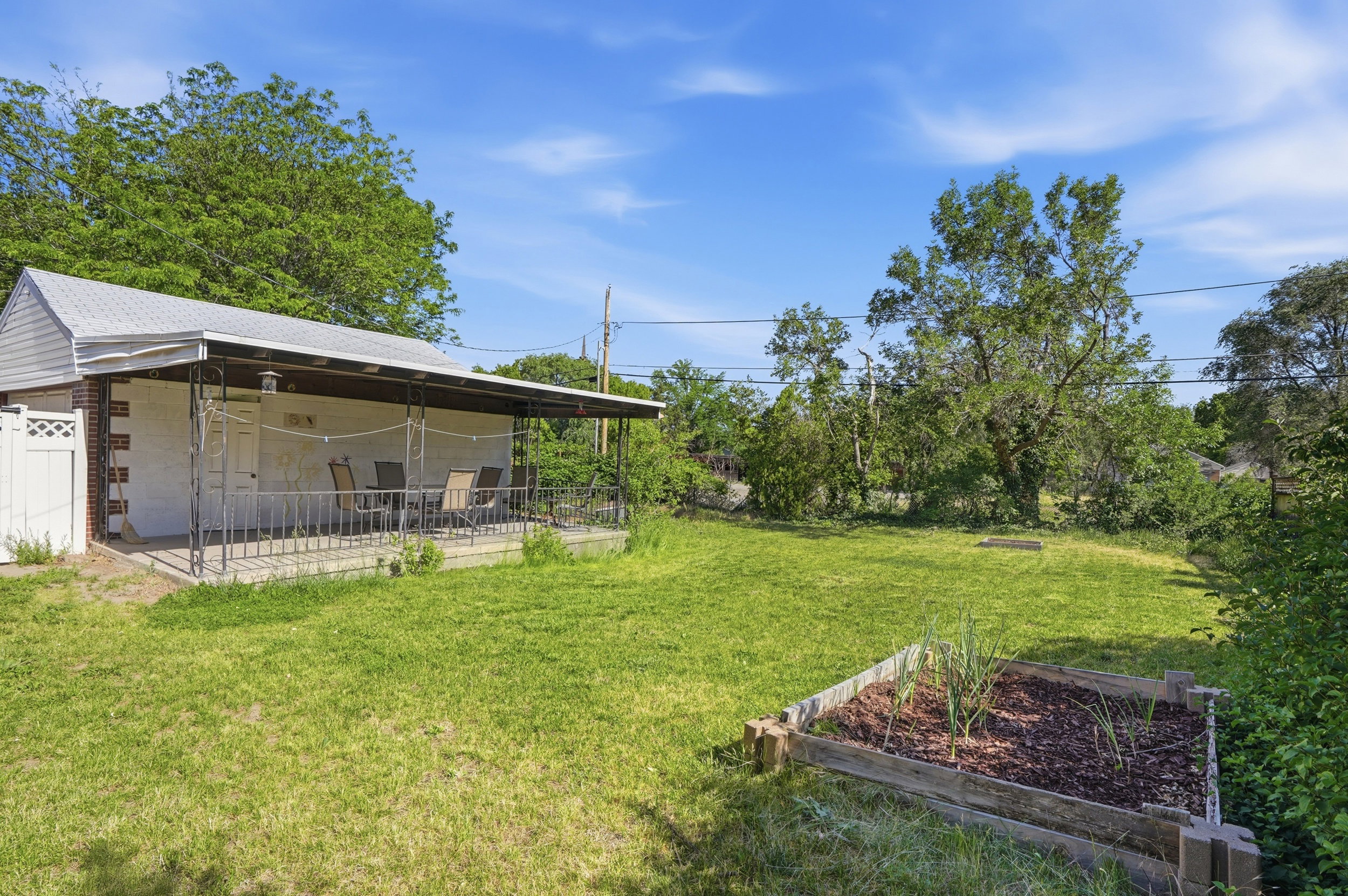 Expansive backyard with a covered patio featuring wrought iron railings, a white privacy fence, and a raised garden bed