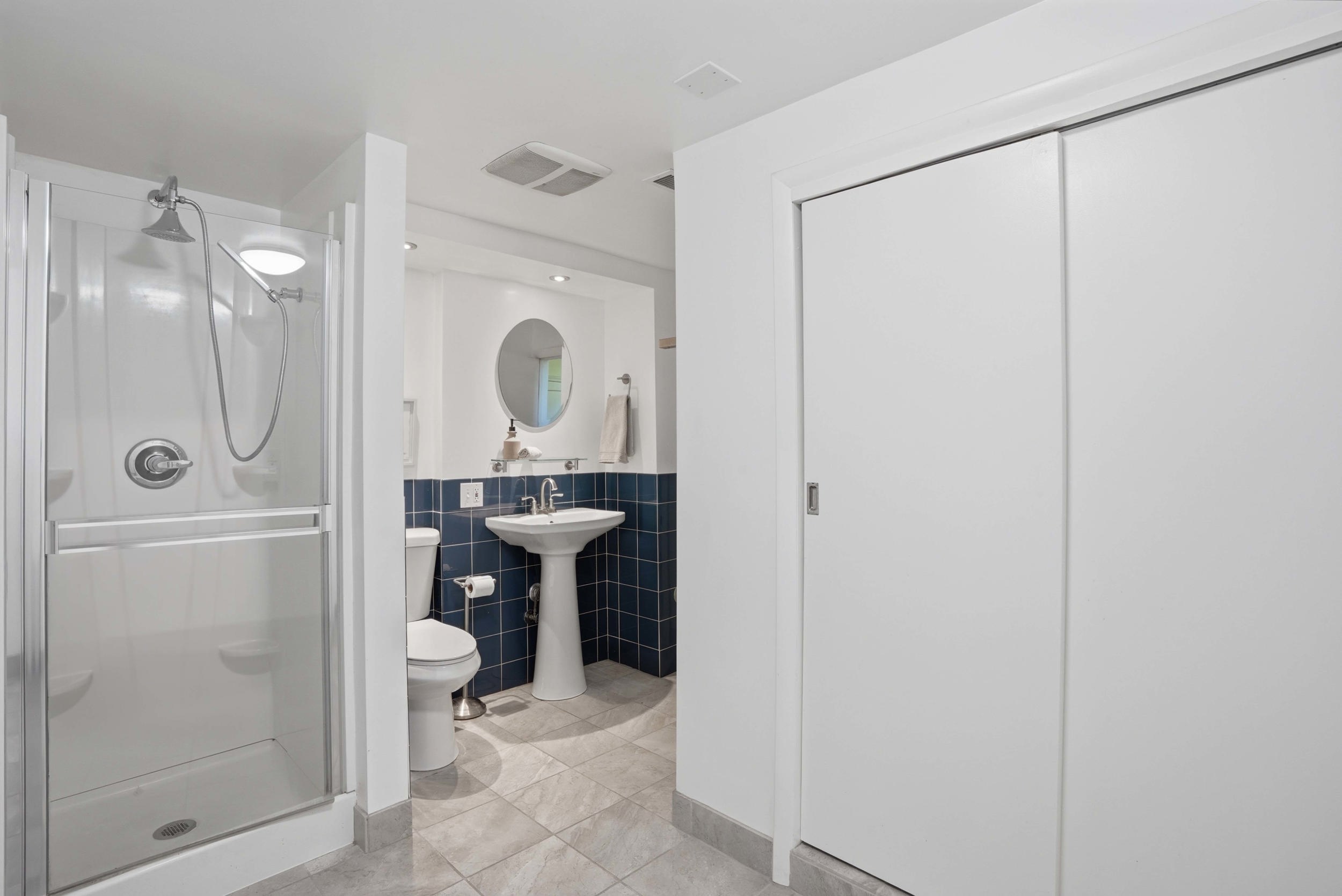 Bathroom featuring a glass-enclosed shower, white pedestal sink, toilet, dark blue square tile accent wall, and light-toned floor tiles