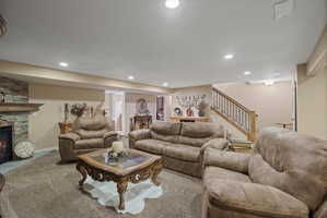 Spacious living area featuring a stacked stone fireplace with a substantial wood mantel, recessed lighting, carpet flooring, and a hardwood staircase with wrought iron balusters