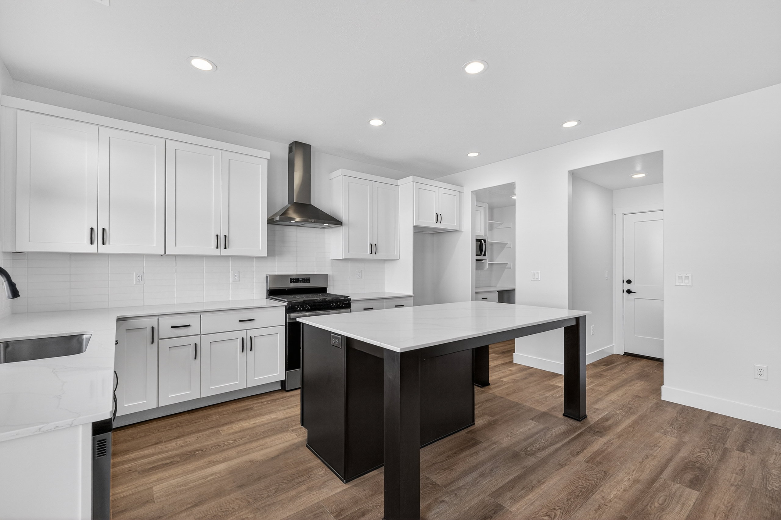 Modern kitchen featuring wood-finish flooring, white cabinetry with black hardware, a stainless steel range with a coordinating hood, an island with a light-colored countertop, and recessed lighting