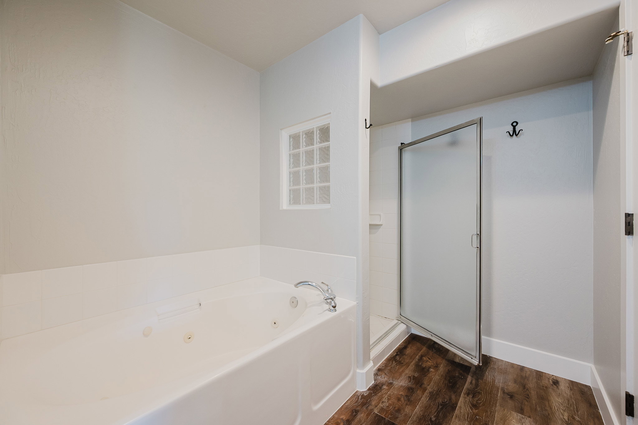 Bathroom featuring a white jetted soaking tub, glass block accent window, walk-in shower with frosted glass door, and wood-finish flooring