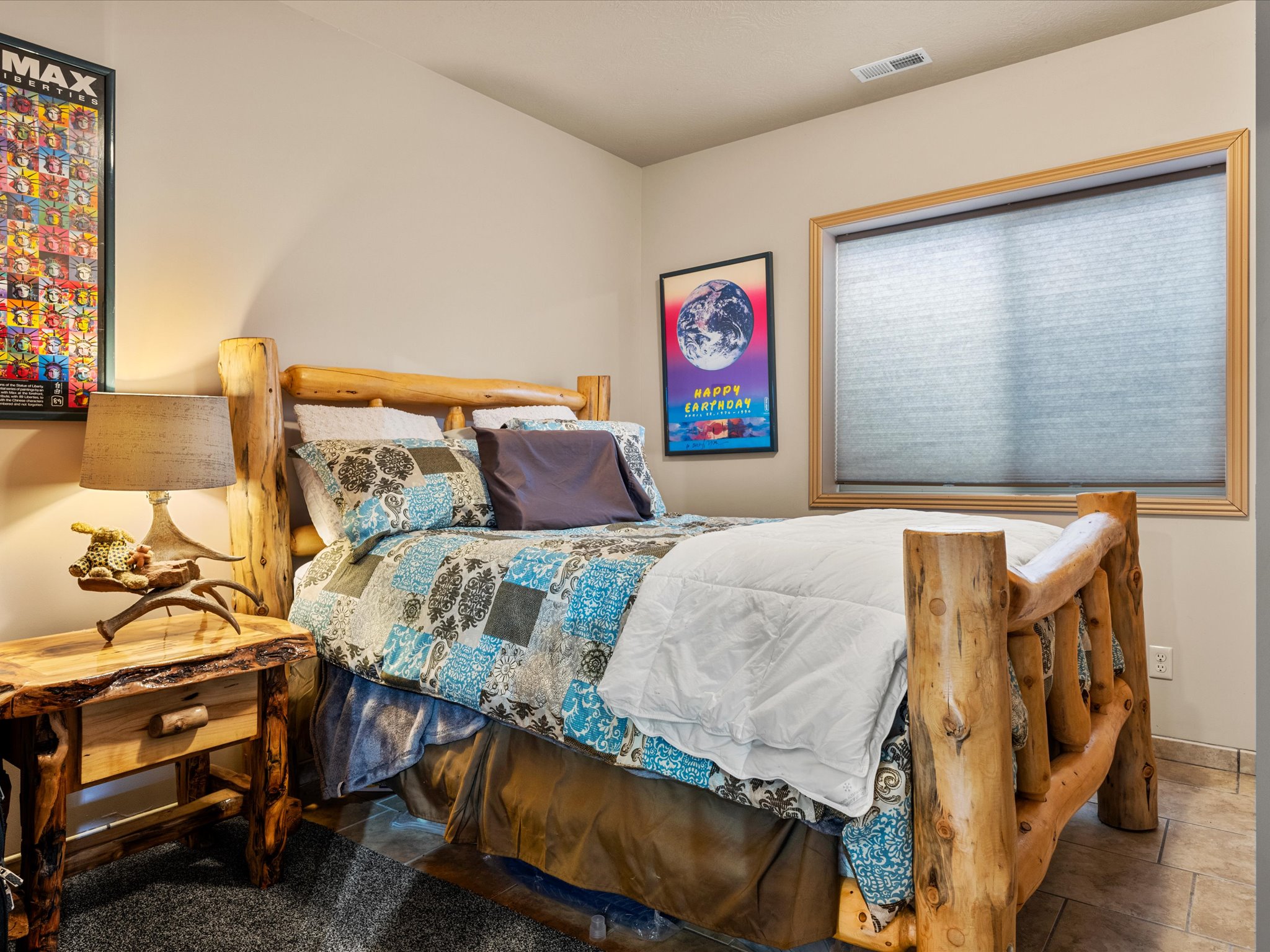 Bedroom featuring a rustic log bed frame, coordinating wood-finish nightstand with drawer, and a single window with wood-finish trim and roller shade