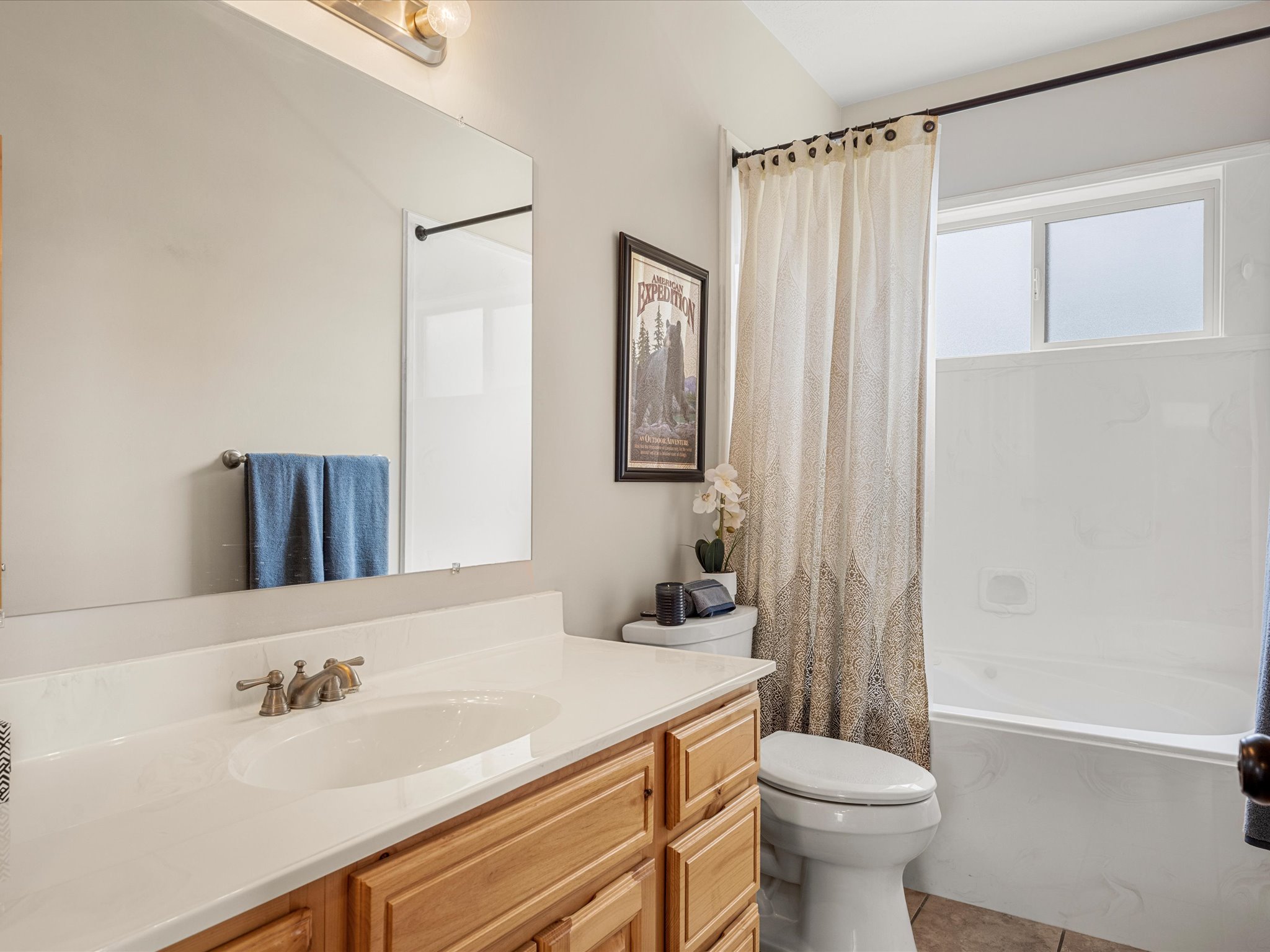 Bathroom featuring an integrated vanity sink with a light-toned countertop, a wood-finish vanity cabinet, a wall-mounted mirror, a toilet, and a combination tub/shower with a window