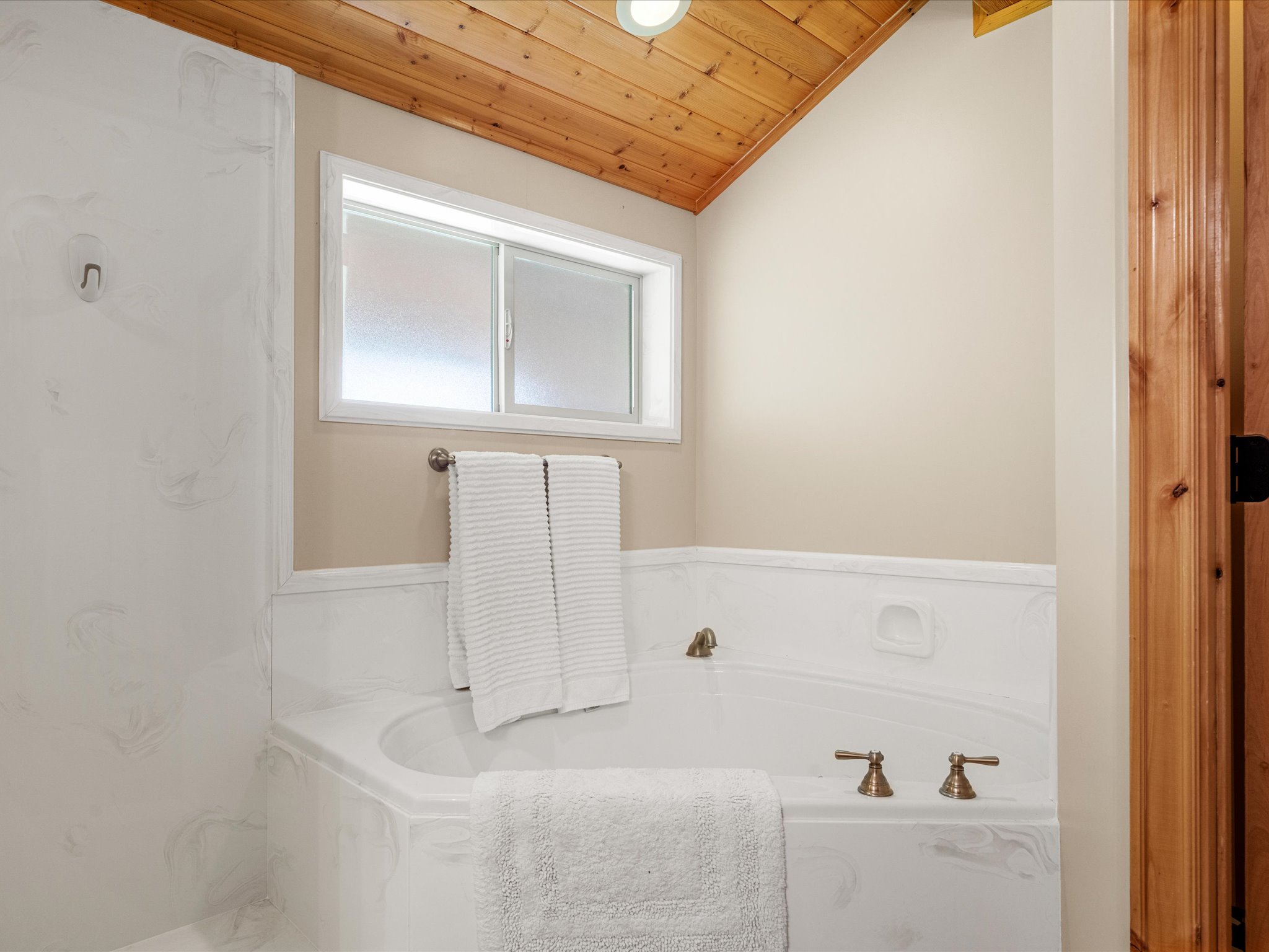 Built-in corner bathtub with brass fixtures, a frosted window, and a wood-paneled vaulted ceiling
