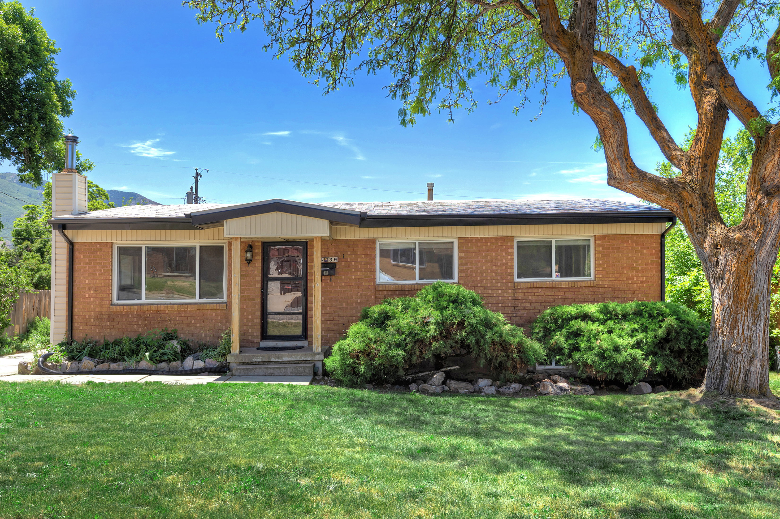 Brick exterior featuring a covered entry, established landscaping, a mature tree, and a manicured lawn