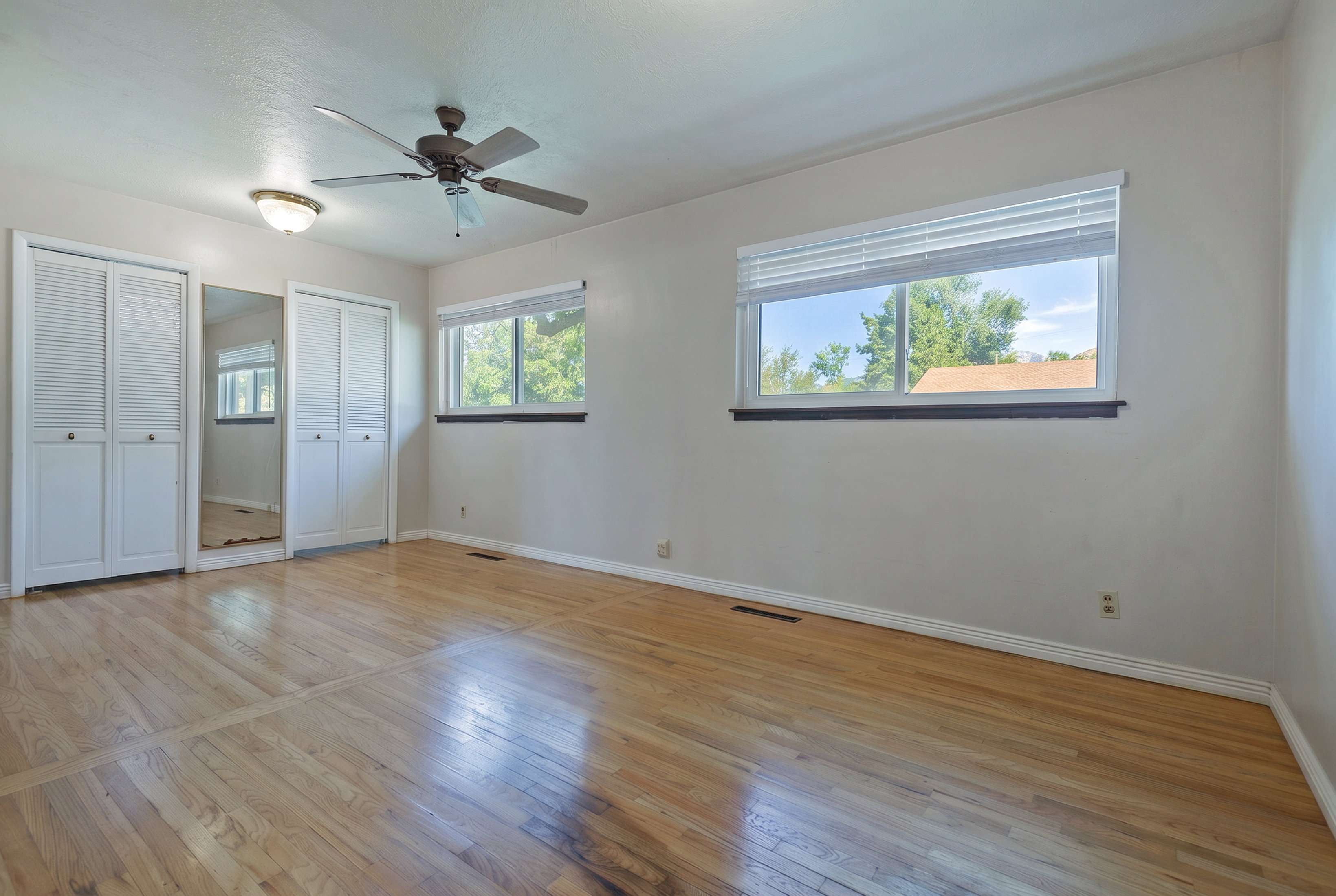 Spacious interior featuring wood-finish flooring, two large windows with blinds, a ceiling fan, and louvered bifold closet doors