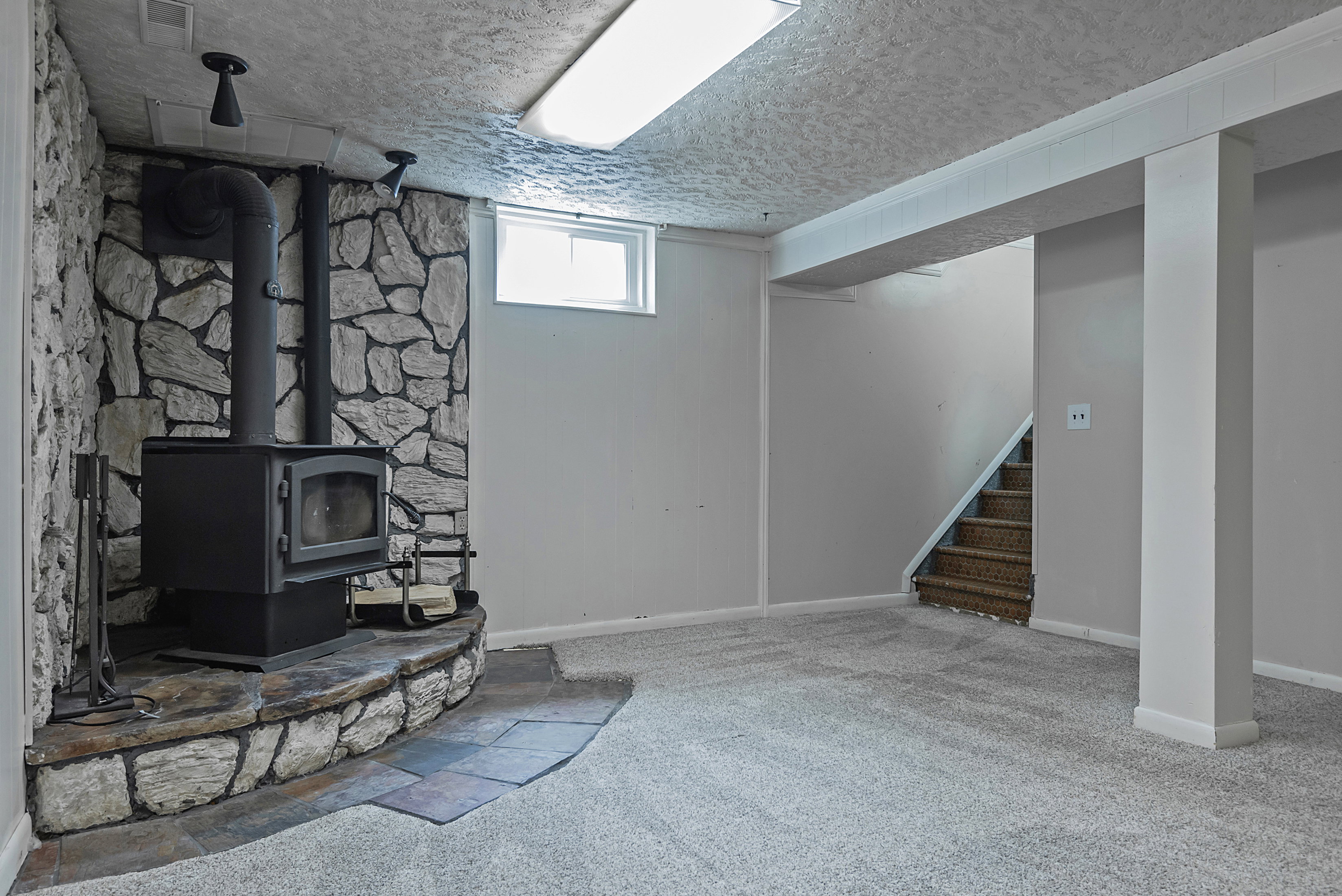 Finished lower level featuring a stone accent wall with a wood-burning stove on a stone hearth, recessed window, and staircase with carpeted treads