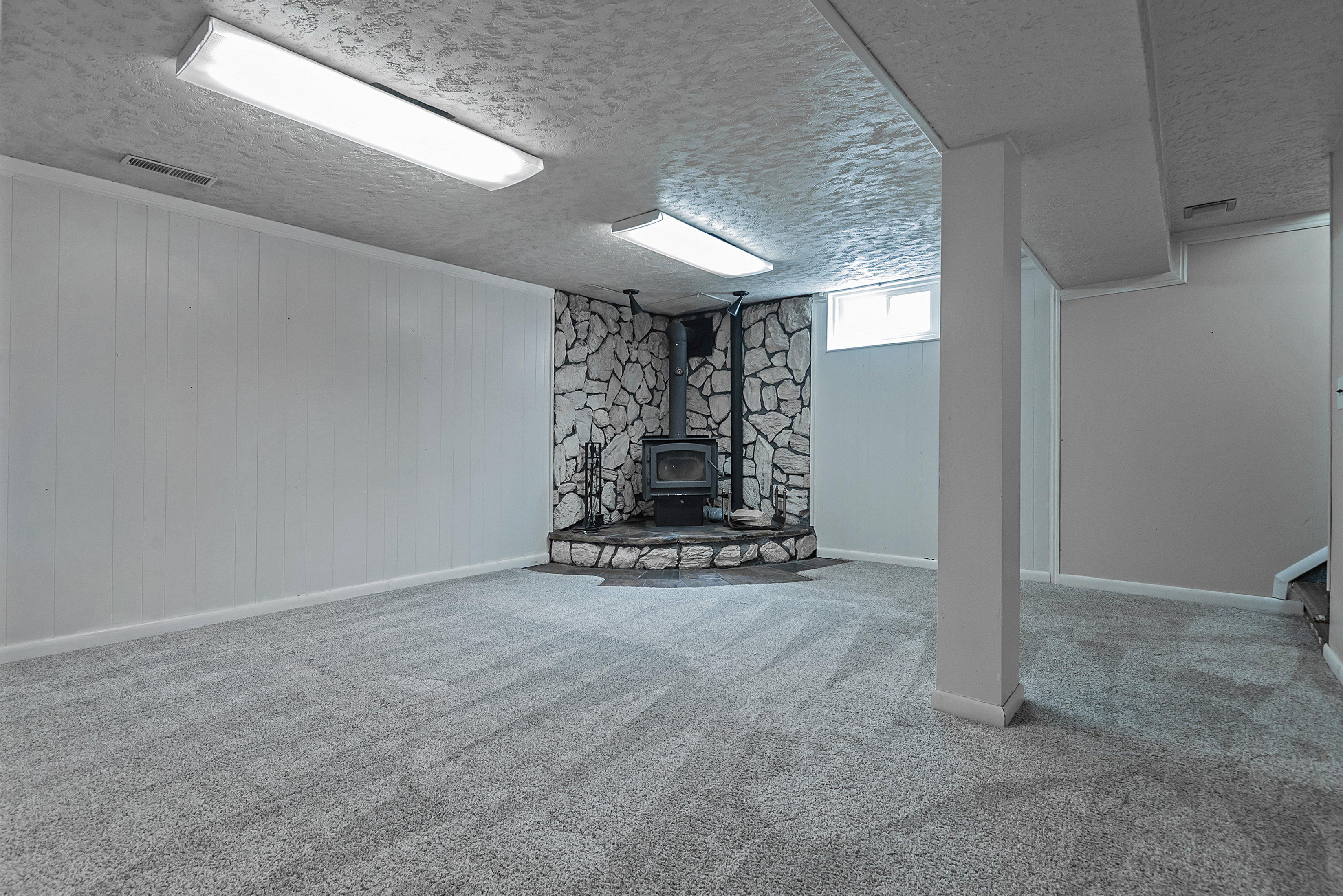Finished lower level featuring a stone fireplace with a wood-burning stove, gray carpet flooring, white paneled walls, and recessed lighting