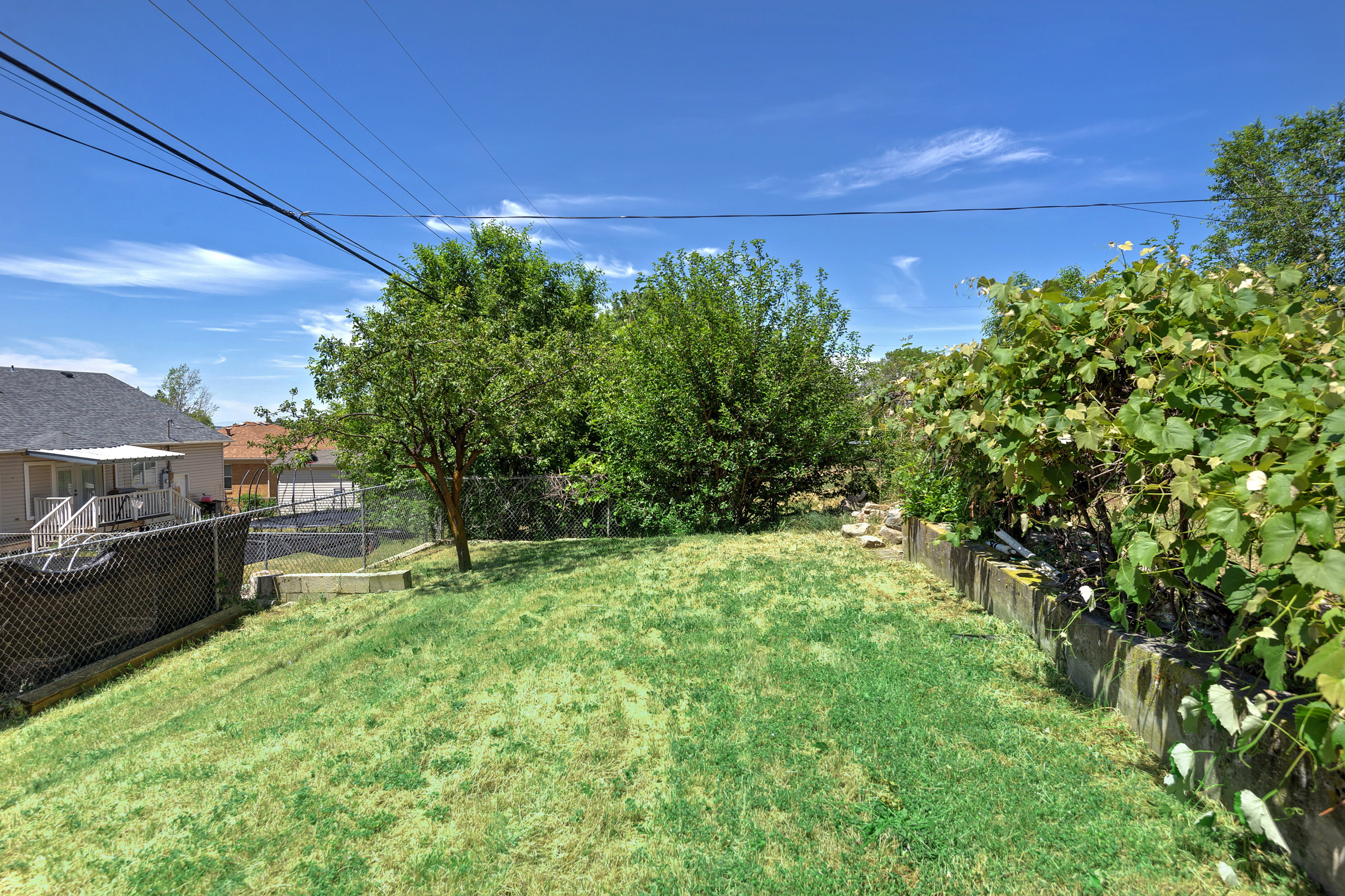 Expansive backyard featuring a grass lawn, mature trees, chain-link fencing, and a concrete block retaining wall with lush green foliage