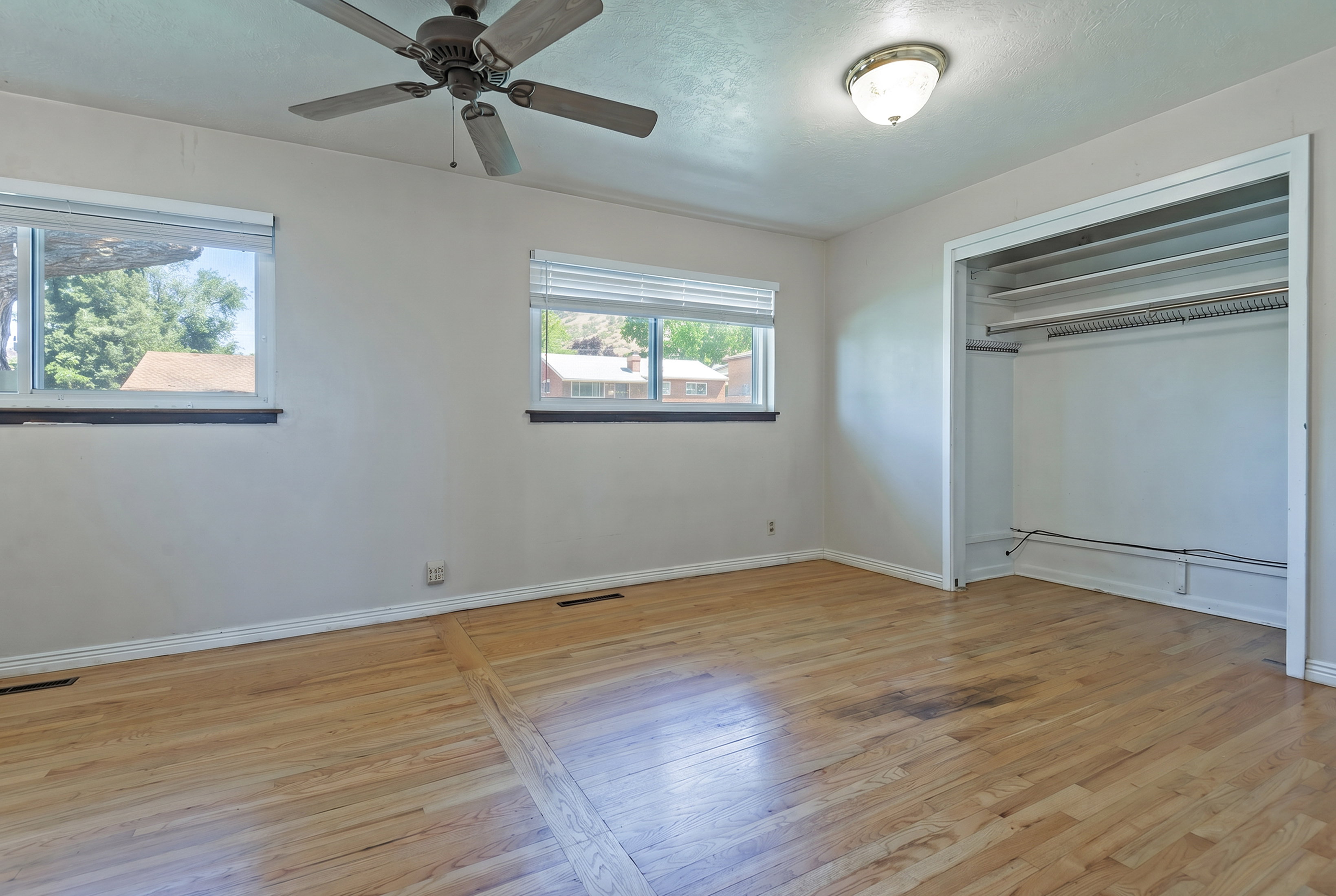 Room featuring wood-finish flooring, two windows with blinds, an open closet with shelving and hanging rod, a ceiling fan, and an overhead light fixture