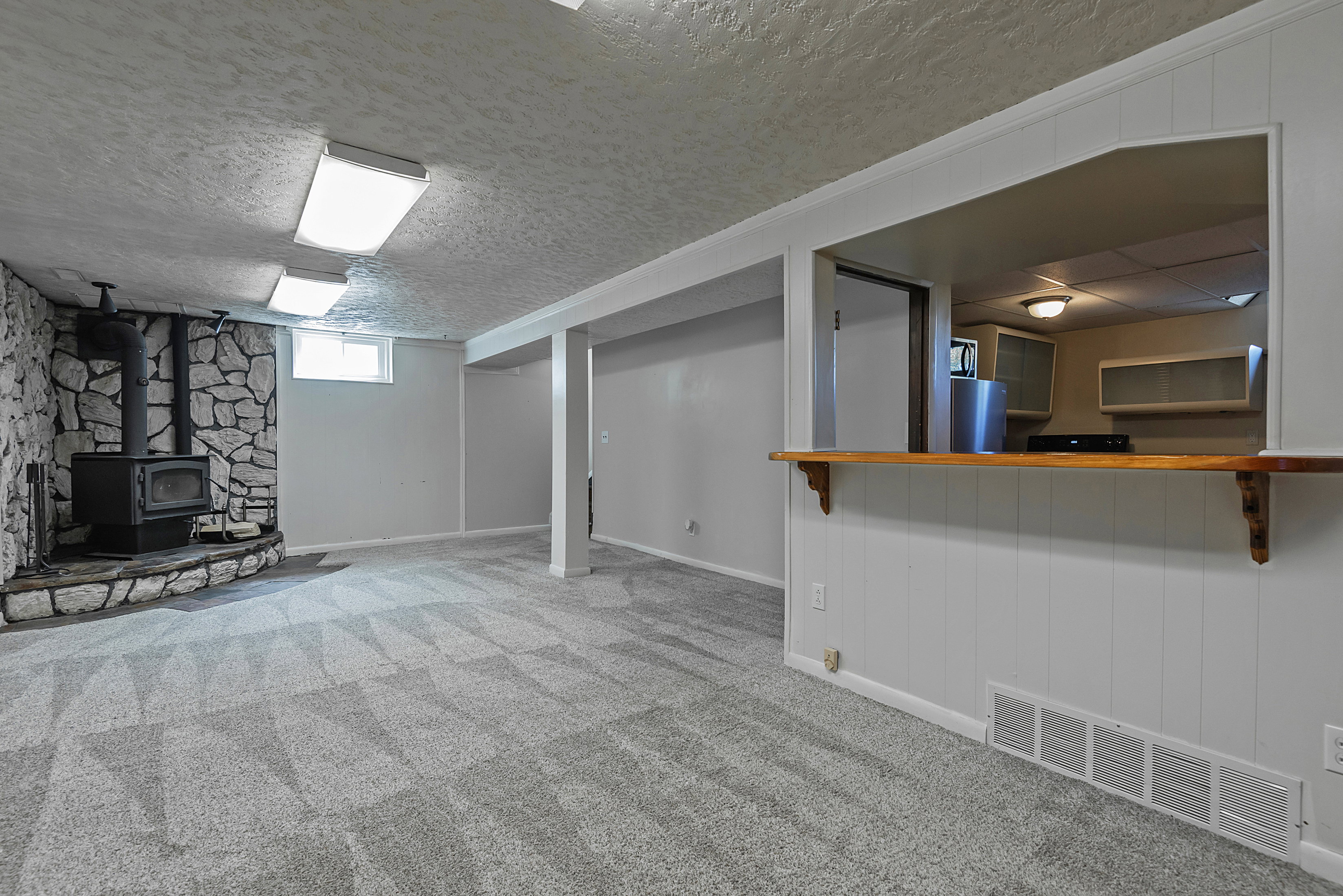Spacious carpeted room featuring a stone-clad wood-burning stove, a window, and overhead lighting