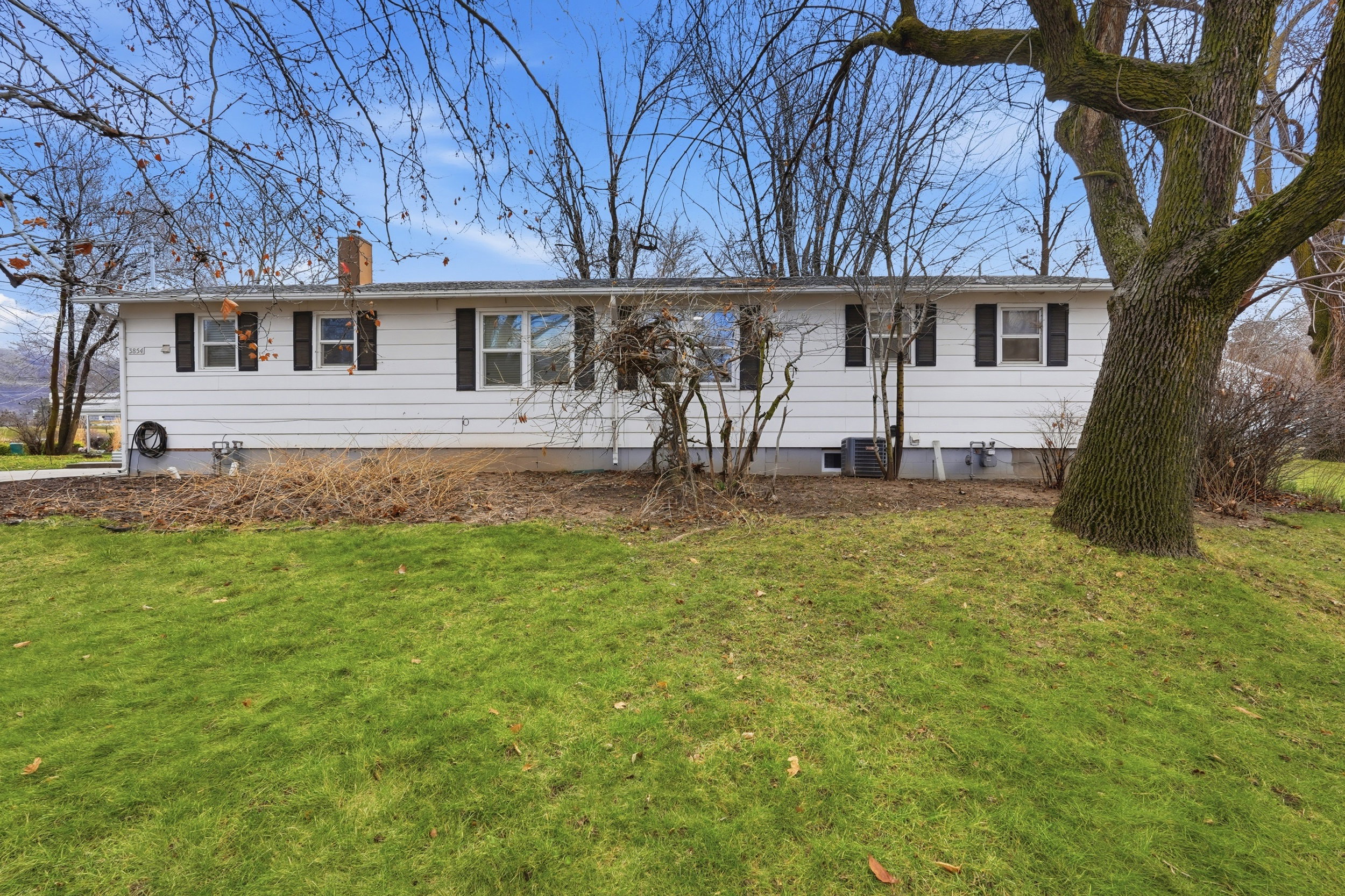 White siding exterior featuring multiple windows with black shutters, a brick chimney, and a sloped grassy lawn