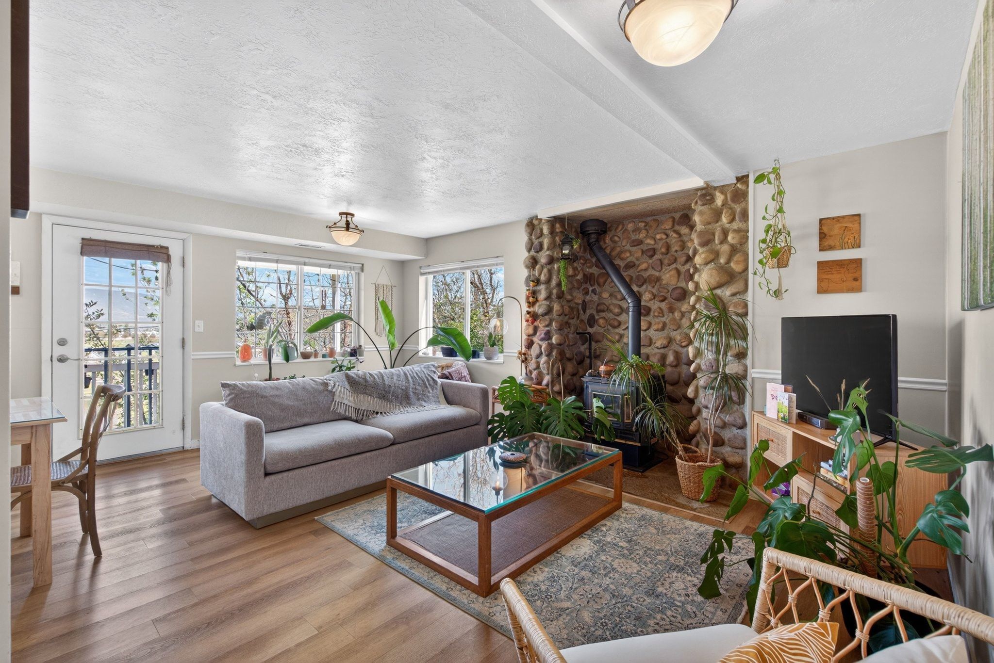 Living area featuring wood-finish flooring, a stone-clad wall with a wood-burning stove, multiple windows, and a glass-paneled door