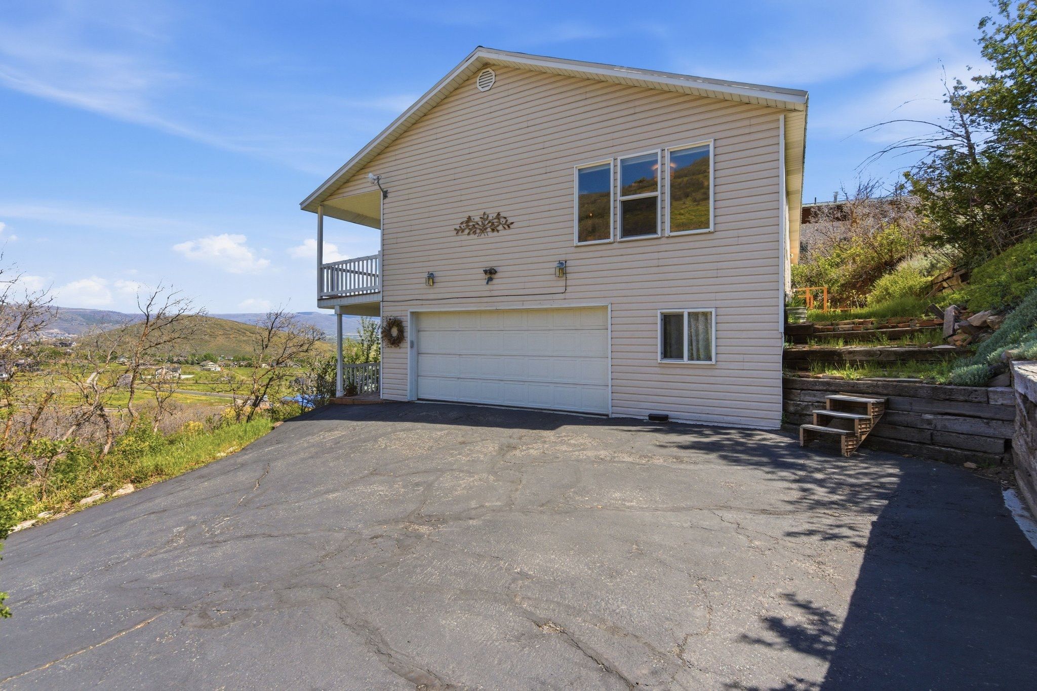 Beige siding exterior featuring a two-car garage, upper-level balcony, paved driveway, and terraced landscaping