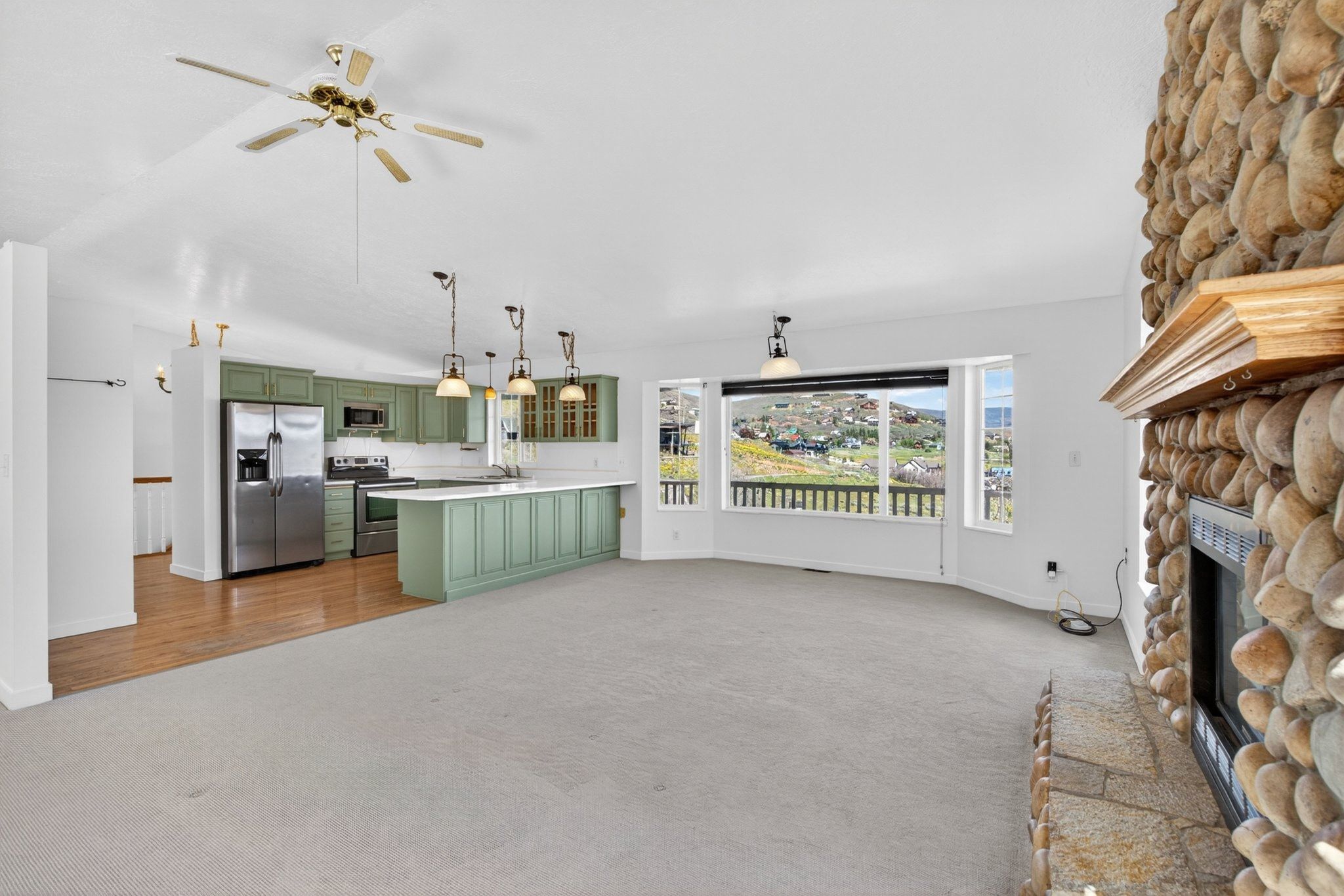 Open-concept living area featuring a stone fireplace with wood mantel, ample natural light from multiple windows, a kitchen with sage green cabinetry and stainless steel appliances, and wood-finish flooring in the kitchen area