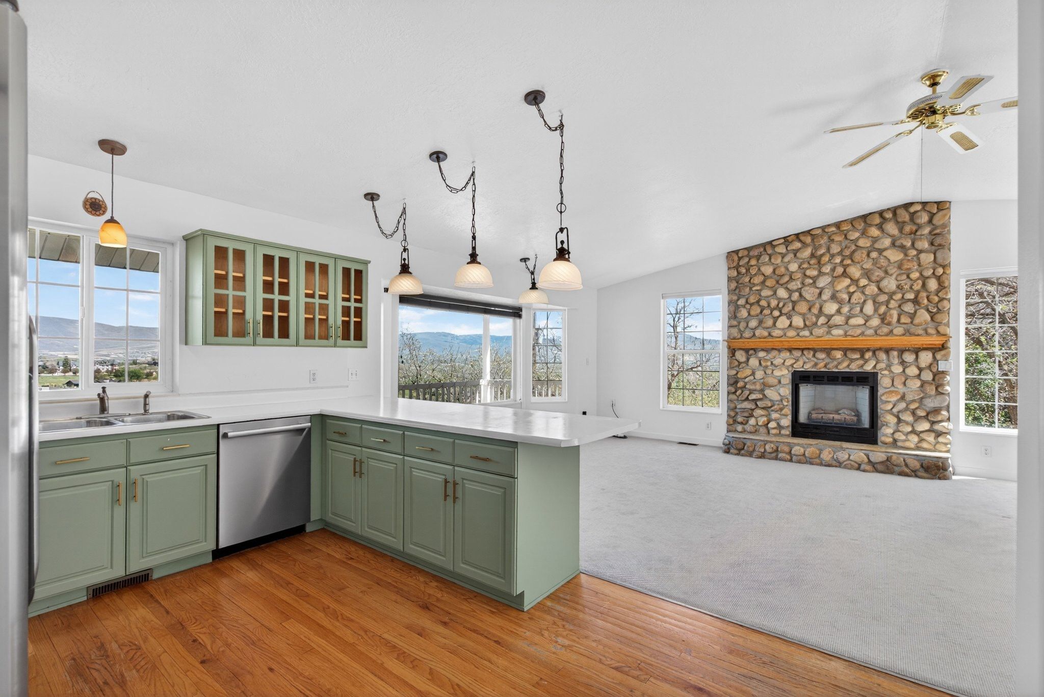 Open-concept living space featuring a spacious kitchen with sage green cabinetry and white countertops, hardwood-finish flooring, a large stone fireplace, and ample natural light from multiple windows