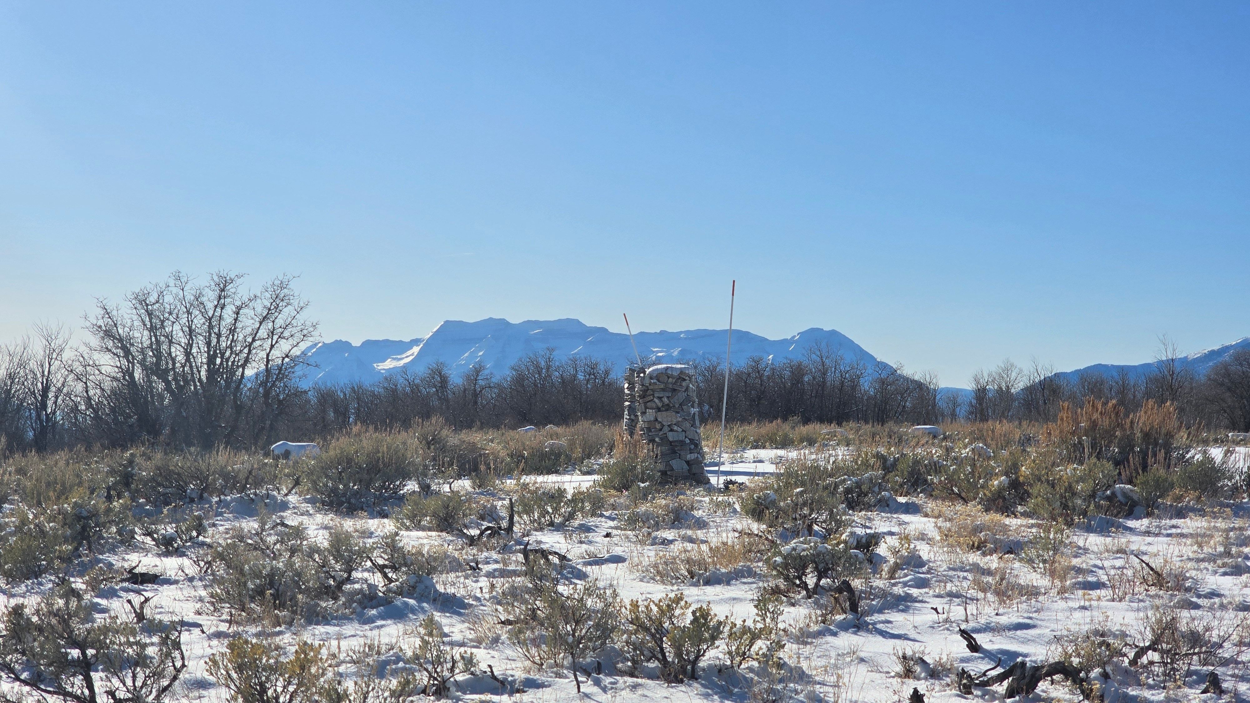 Expansive undeveloped land featuring a partial stone column, snow-dusted natural vegetation, and a clear mountain backdrop under a bright blue sky