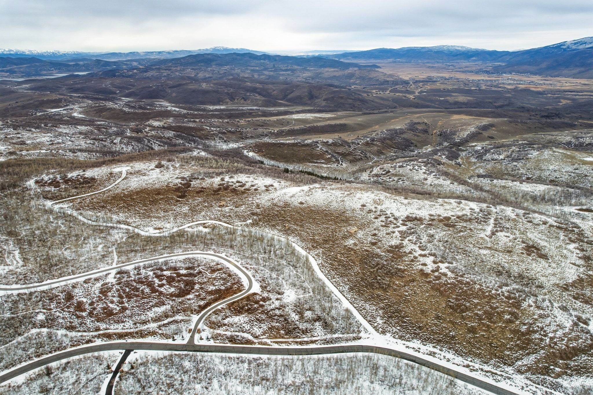 Expansive aerial landscape featuring a mountainous terrain with distant peaks, winding roads, and scattered tree cover