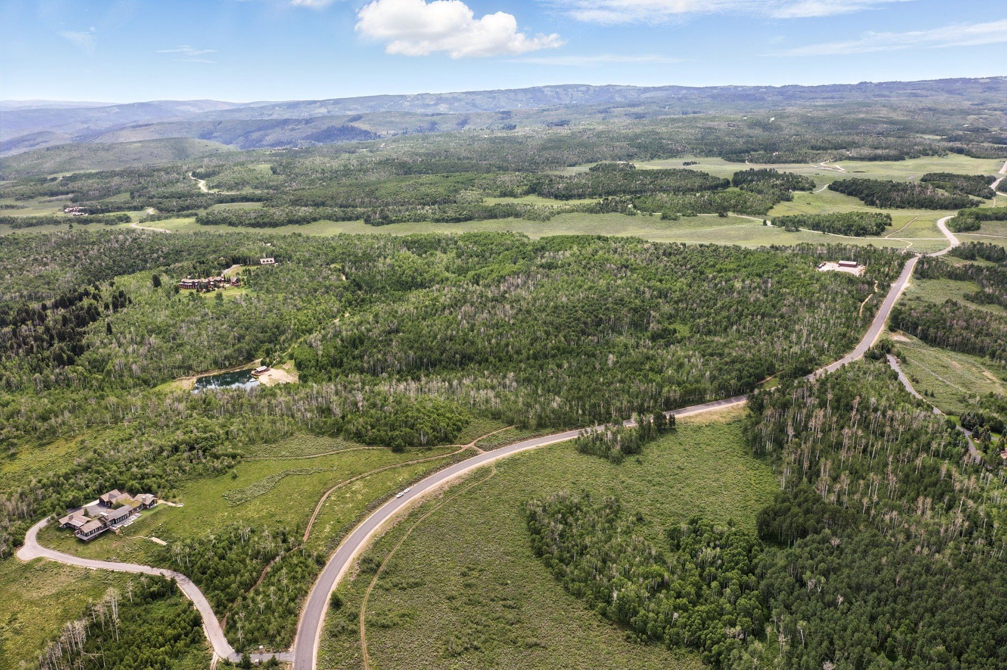 Expansive aerial landscape featuring a winding paved road, lush green tree coverage, distant mountainous terrain, a visible body of water, and residential structures