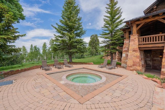 Circular paver patio featuring a built-in spa, surrounding landscaping, and a rustic log and stone exterior