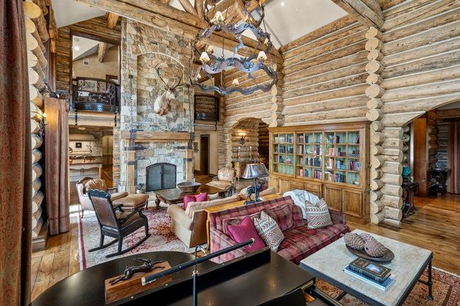 Spacious room featuring log wall construction, a stone-faced fireplace with a hearth, exposed wooden ceiling beams, and polished wood flooring