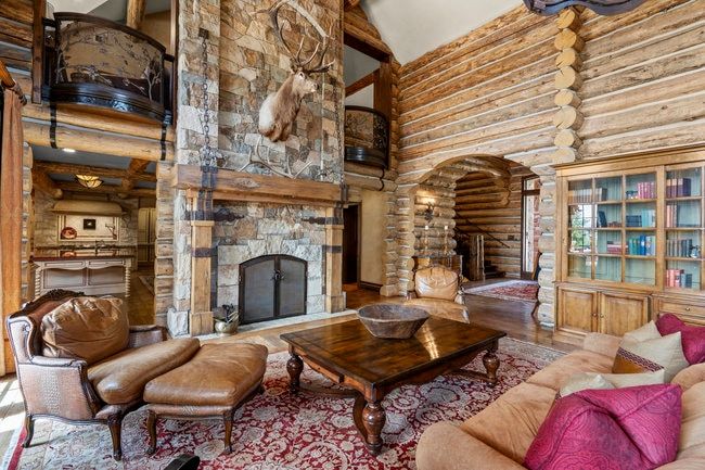 Spacious living area featuring a floor-to-ceiling stone fireplace, exposed log walls, a built-in wood bookcase with glass-front cabinets, a log archway, and a second-story interior balcony with decorative metal railings